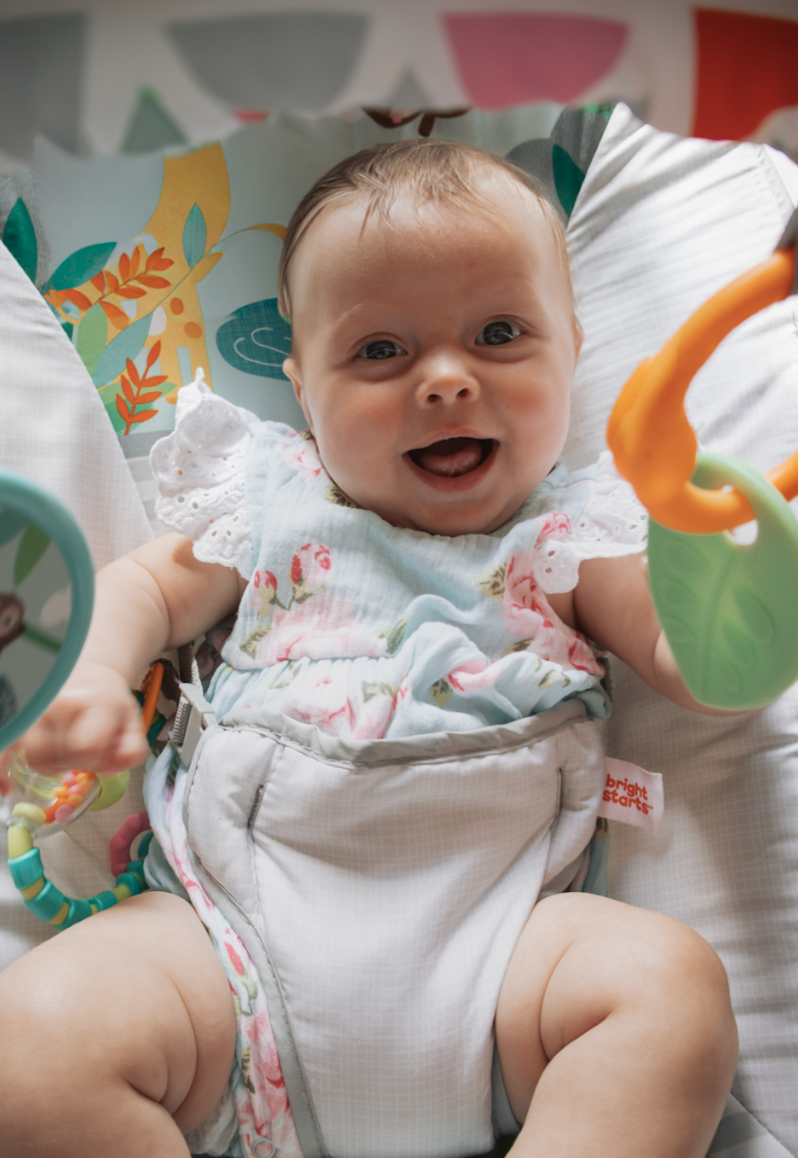 Baby wearing white floral outfit lies on colorful floral bedding while smiling and moving.