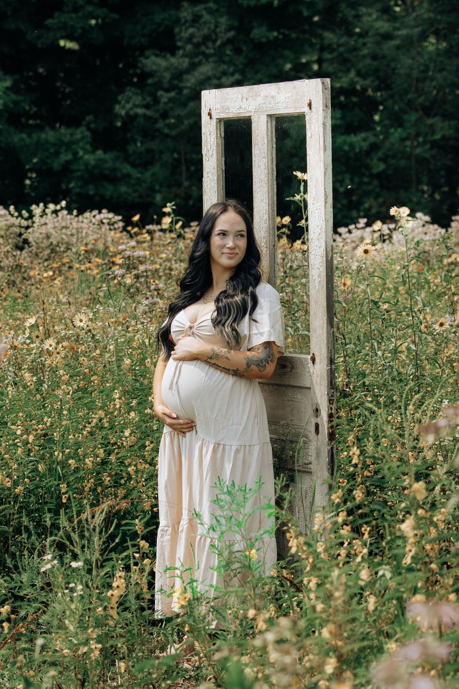 Pregnant woman in white dress standing in wildflower field beside vintage door frame, cradling baby bump.