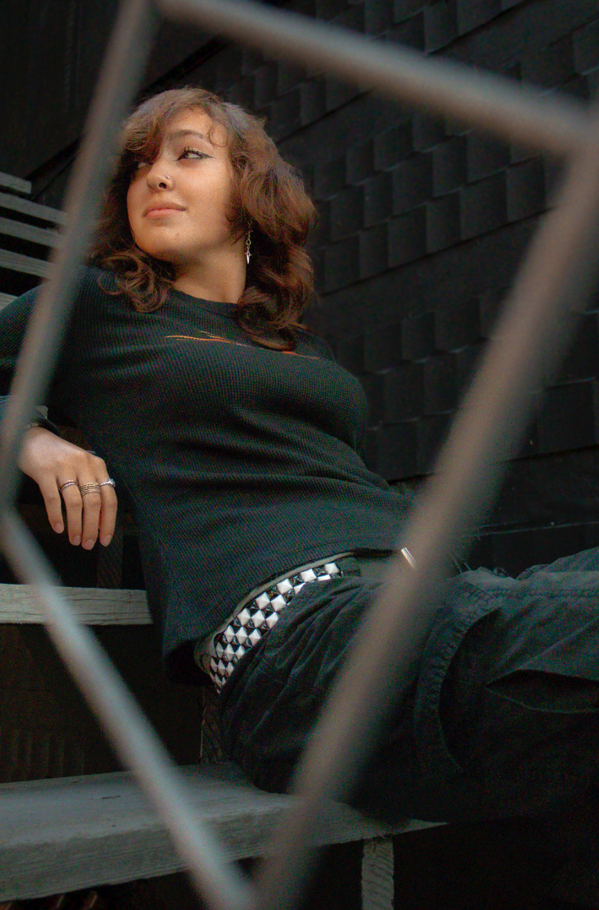 A moody urban photoshoot featuring someone in black clothing against industrial metal bars and dark background.