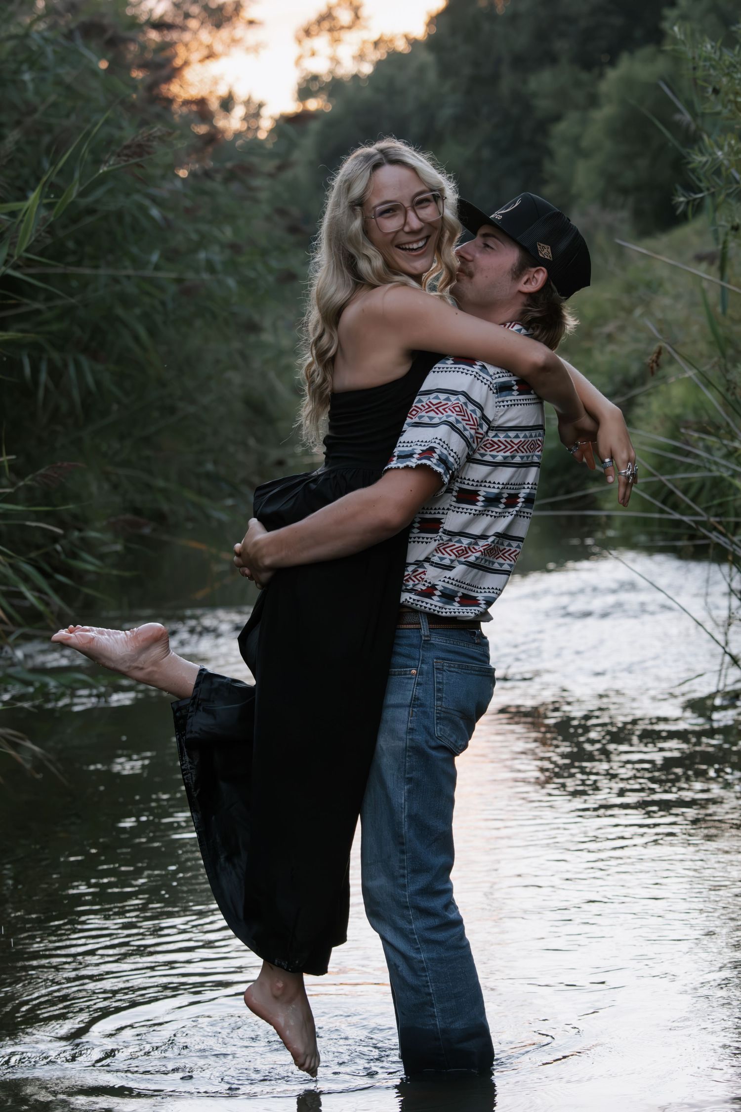 A man lifts and embraces a laughing blonde woman in a black dress while standing in a shallow river surrounded by trees.