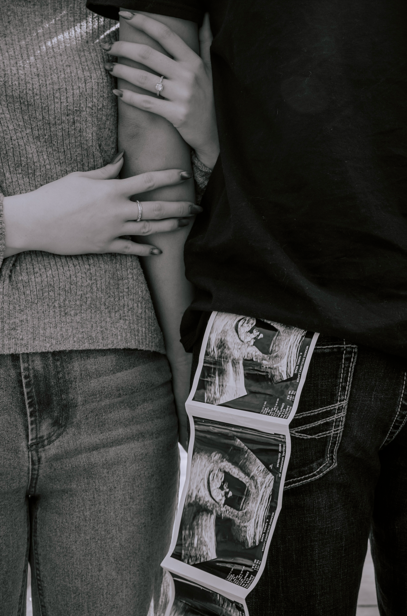 Black and white photo of couple embracing, woman wearing ring, man holding ultrasound photos in back pocket, pregnancy announcement.