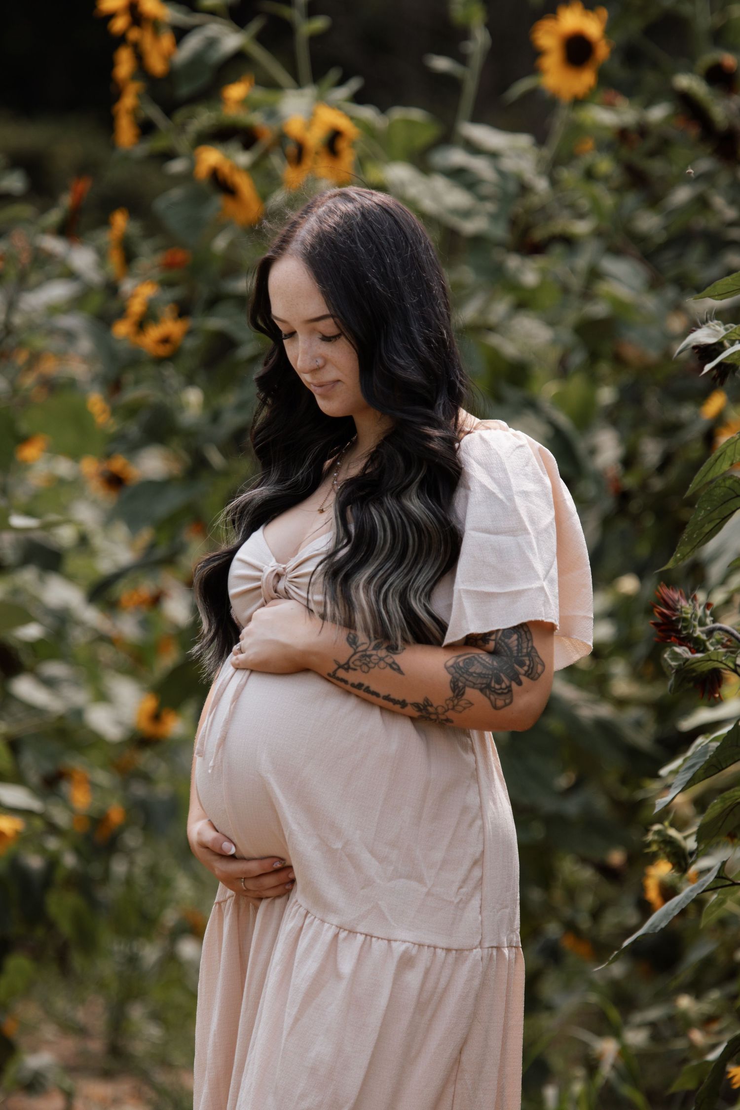 Pregnant woman in blush dress holding baby bump among sunflowers, featuring tattoos and long dark wavy hair.