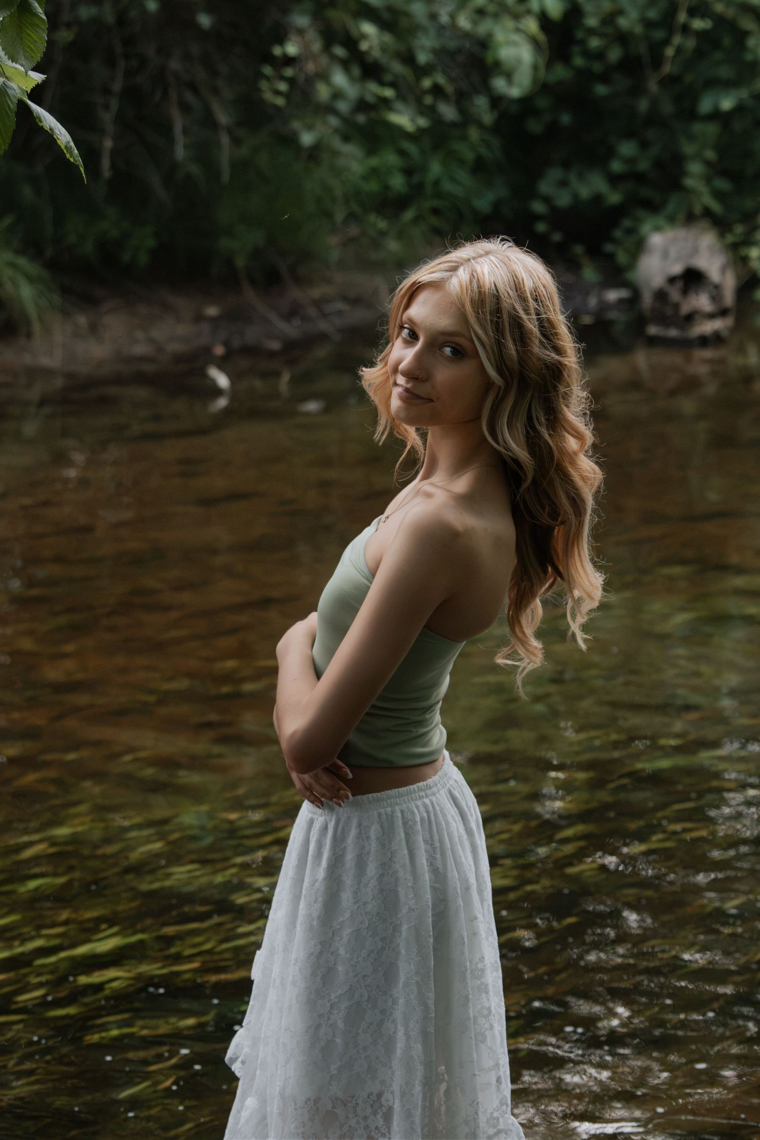 Blonde woman in green crop top and white lace skirt standing in a forest creek.