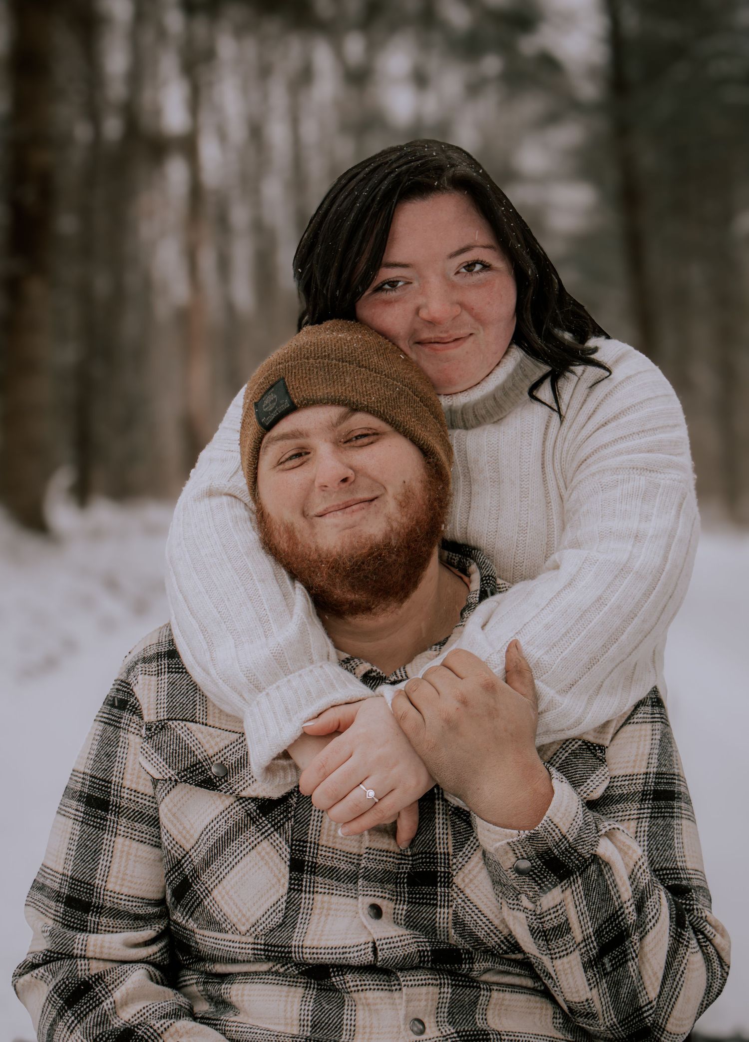 A couple in winter attire shares playful moments in a snowy forest setting during an outdoor photo session.