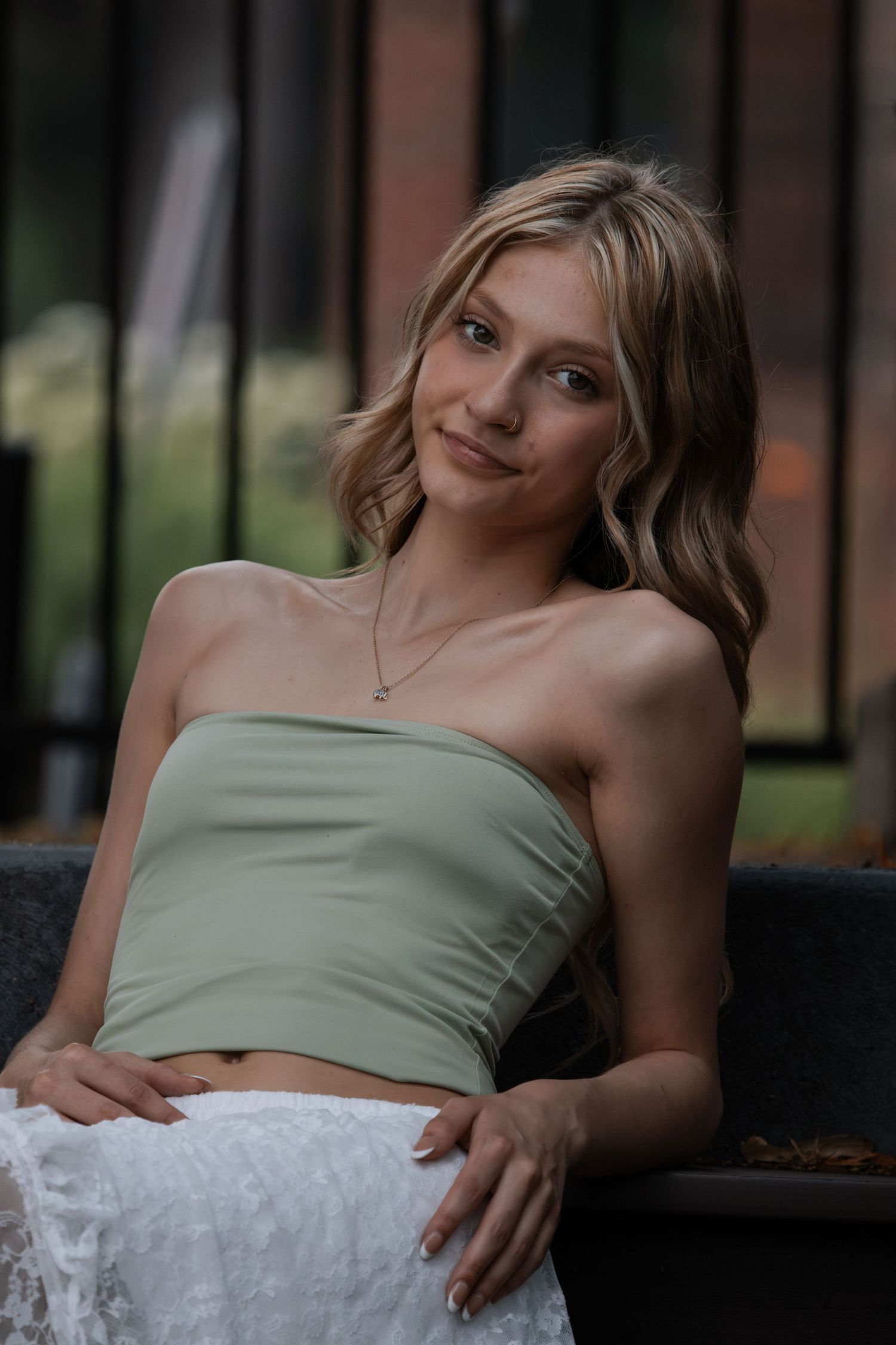 Smiling blonde woman in sage green bandeau top and white skirt seated outdoors.