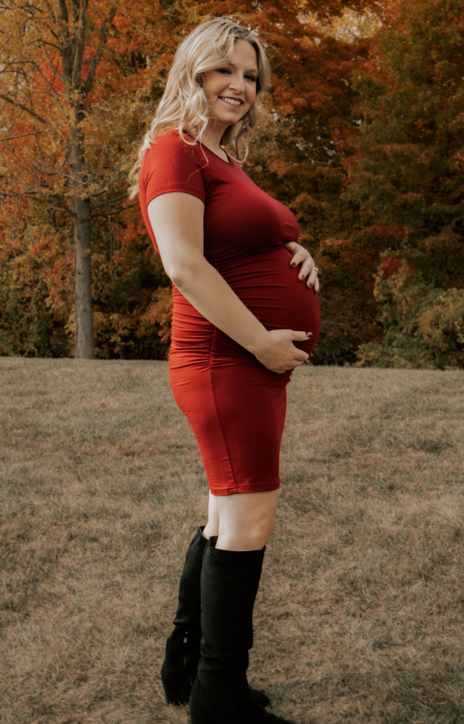 A maternity photoshoot featuring a red dress against a backdrop of fall foliage.