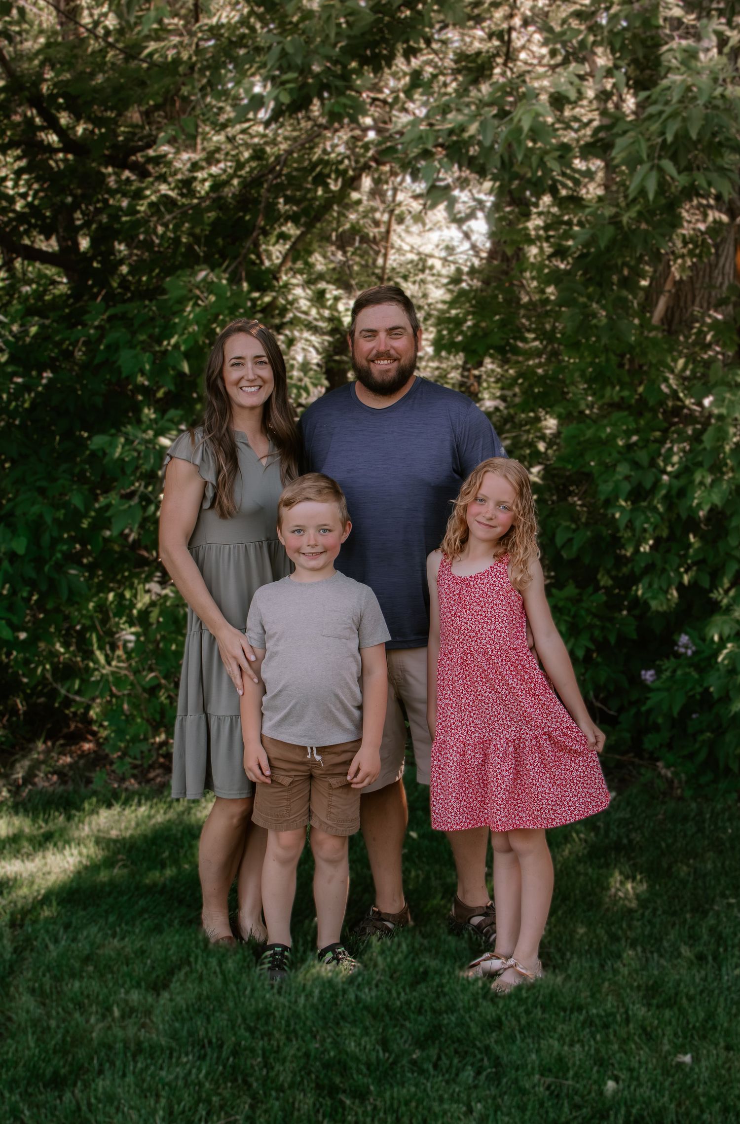 Family of four smiling outdoors on green grass surrounded by lush trees, featuring two adults and two young children.