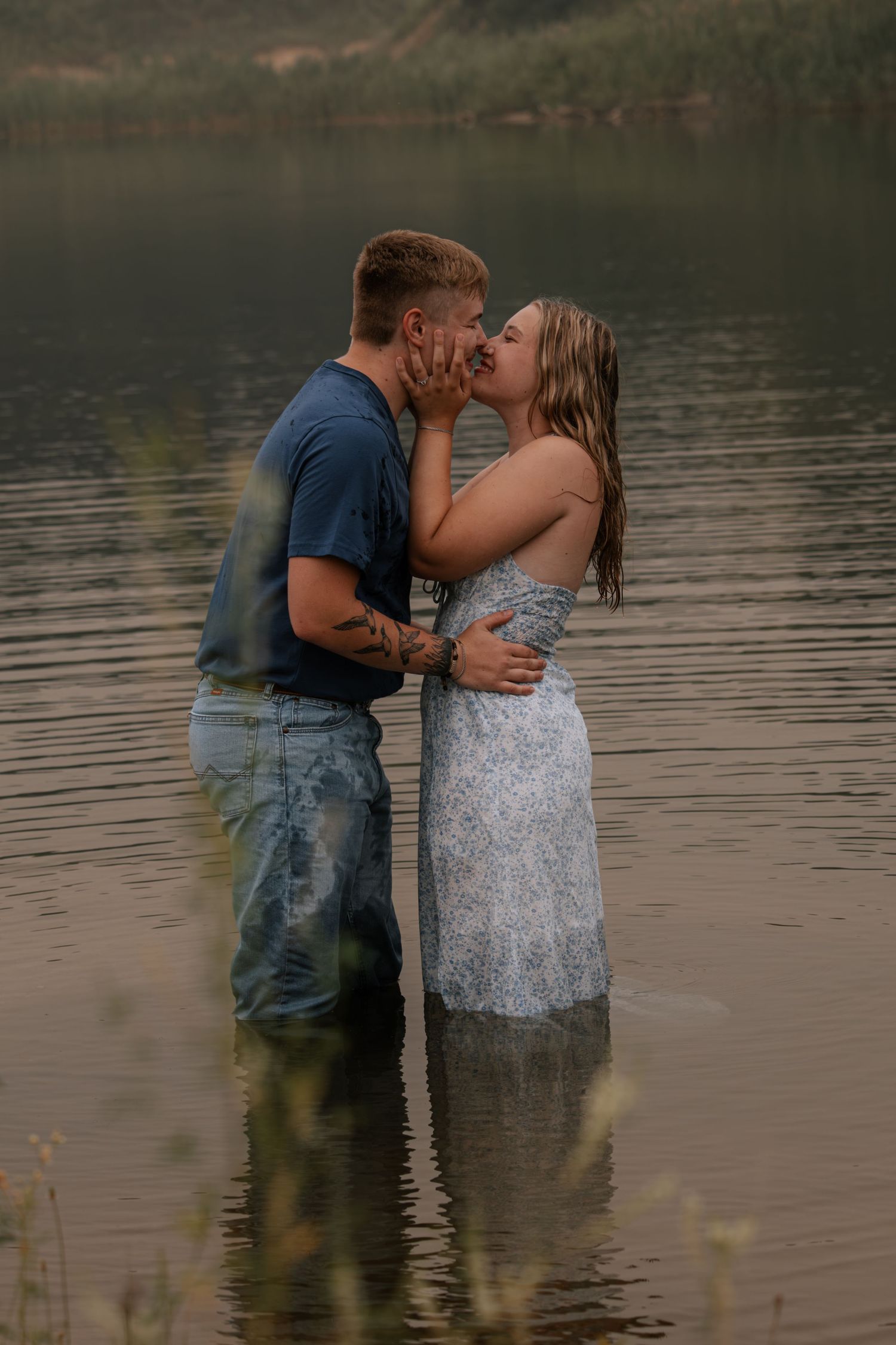 Couple kissing romantically while standing in a lake, man in blue shirt and jeans, woman in white floral dress.