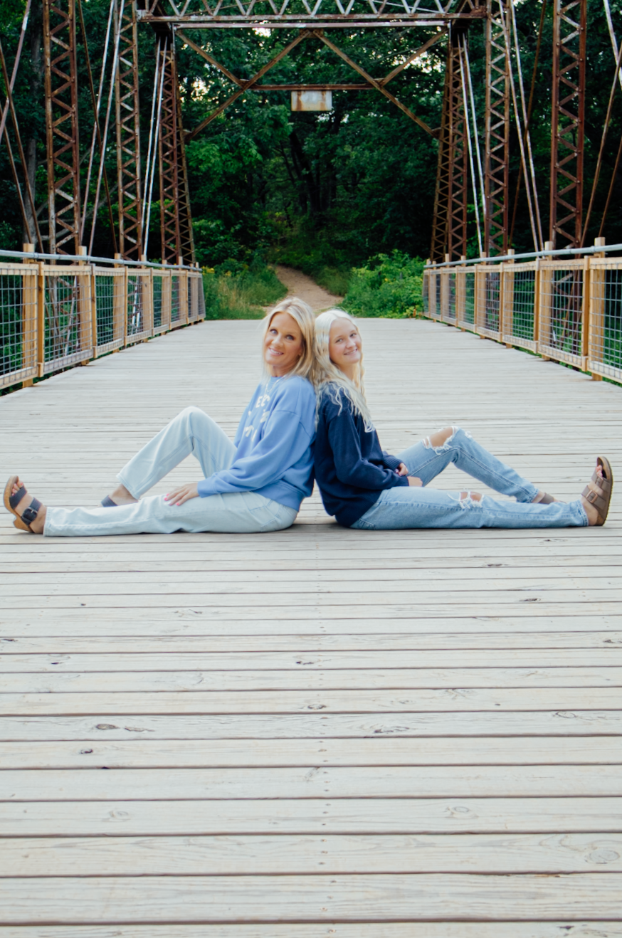 Two people lay casually on wooden bridge planks wearing casual clothing in a natural outdoor setting.