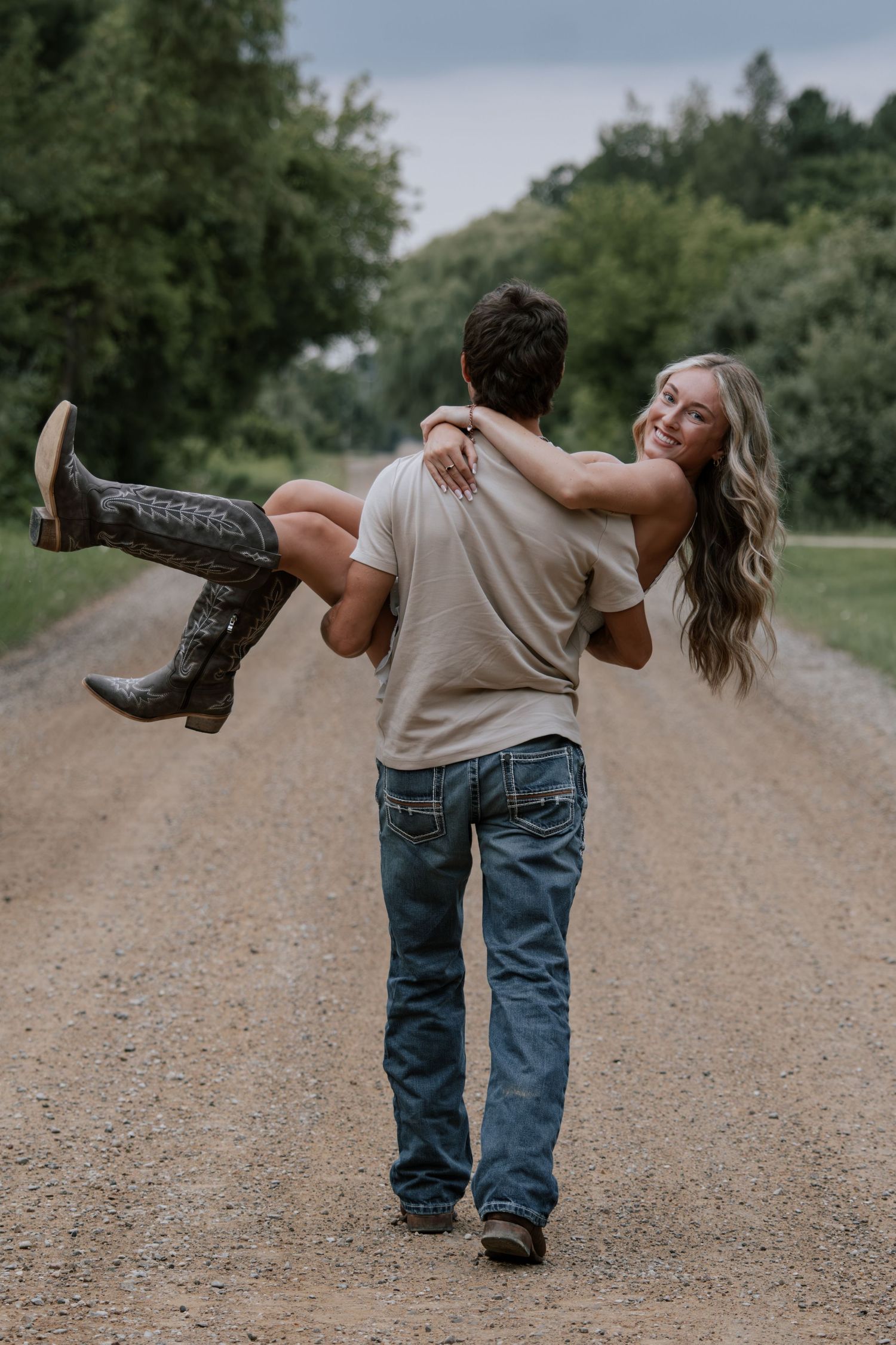 A man carries a smiling woman in cowboy boots down a rural dirt road lined with green trees.
