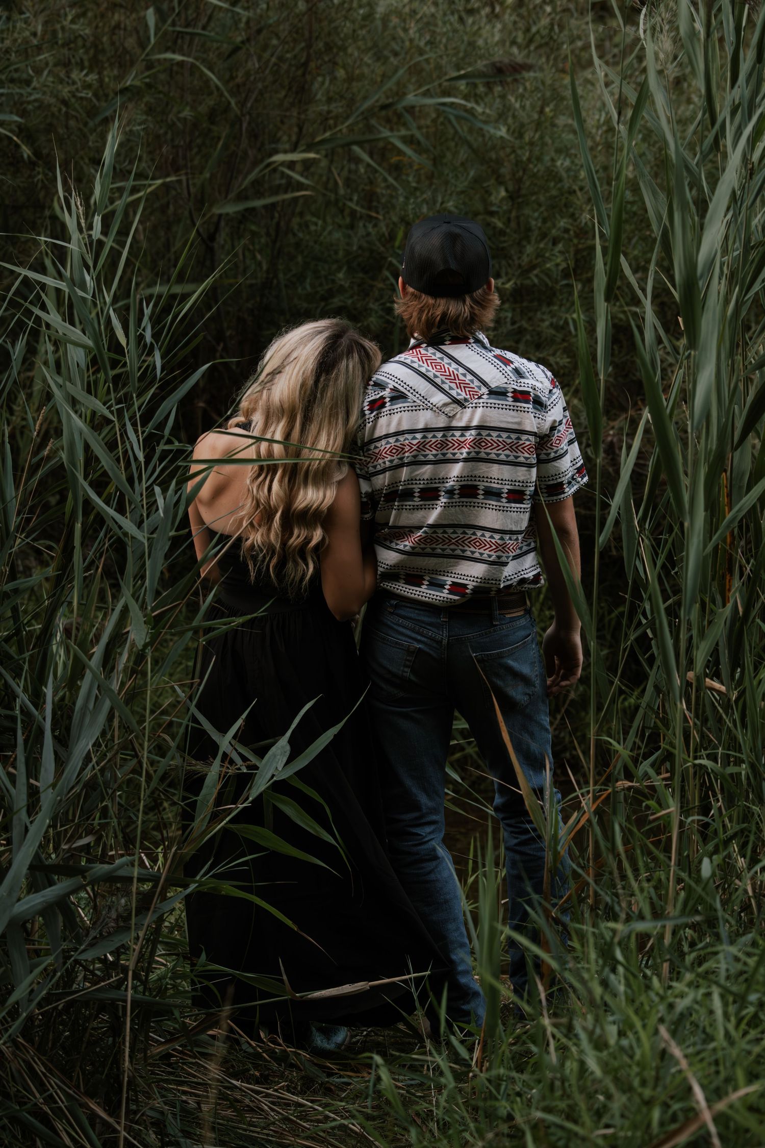 A couple walks hand in hand through tall green reeds, woman in black dress, man in patterned shirt and jeans.