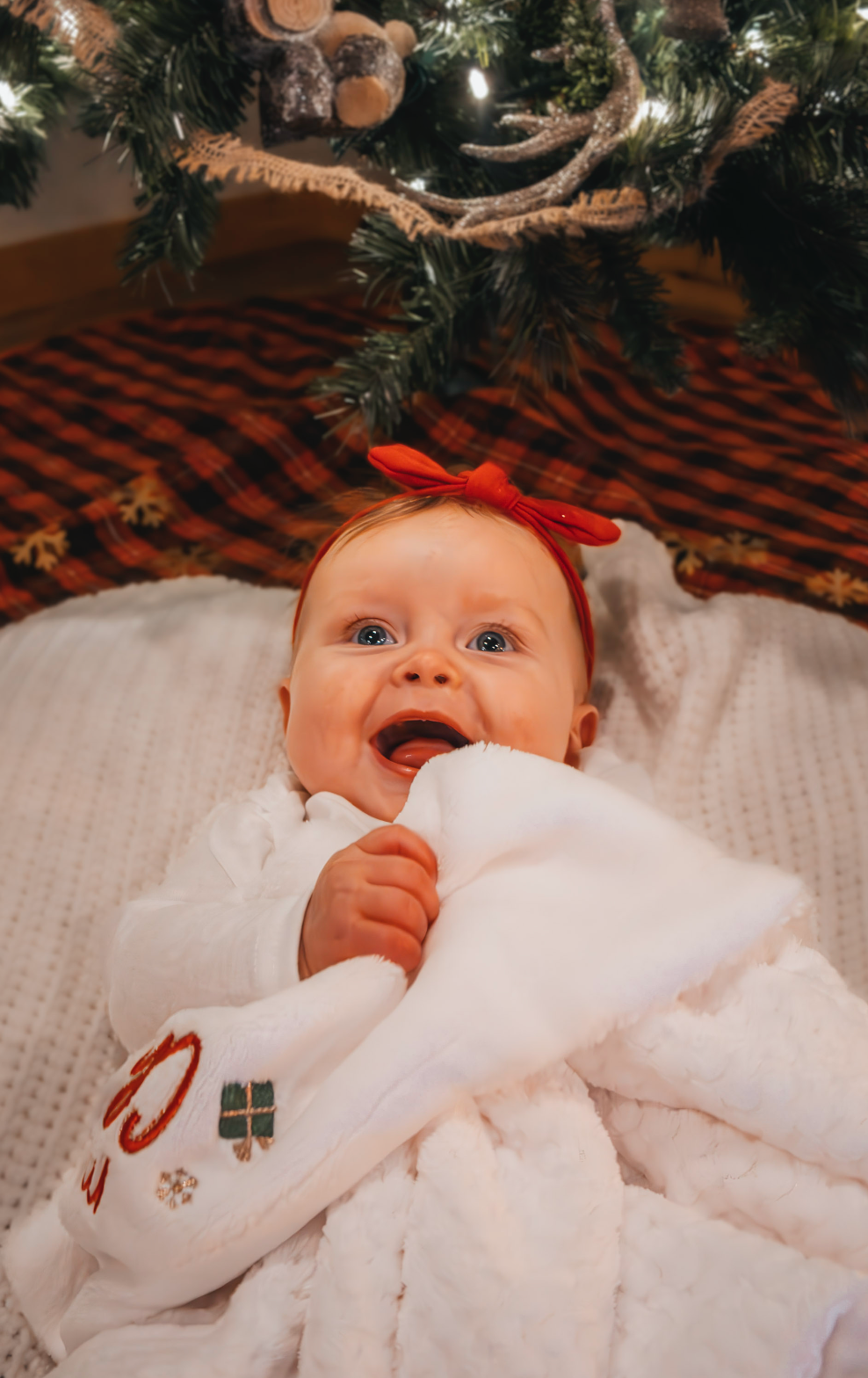 Baby in white outfit with red bow headband lying on blanket under Christmas tree with red and black plaid skirt.