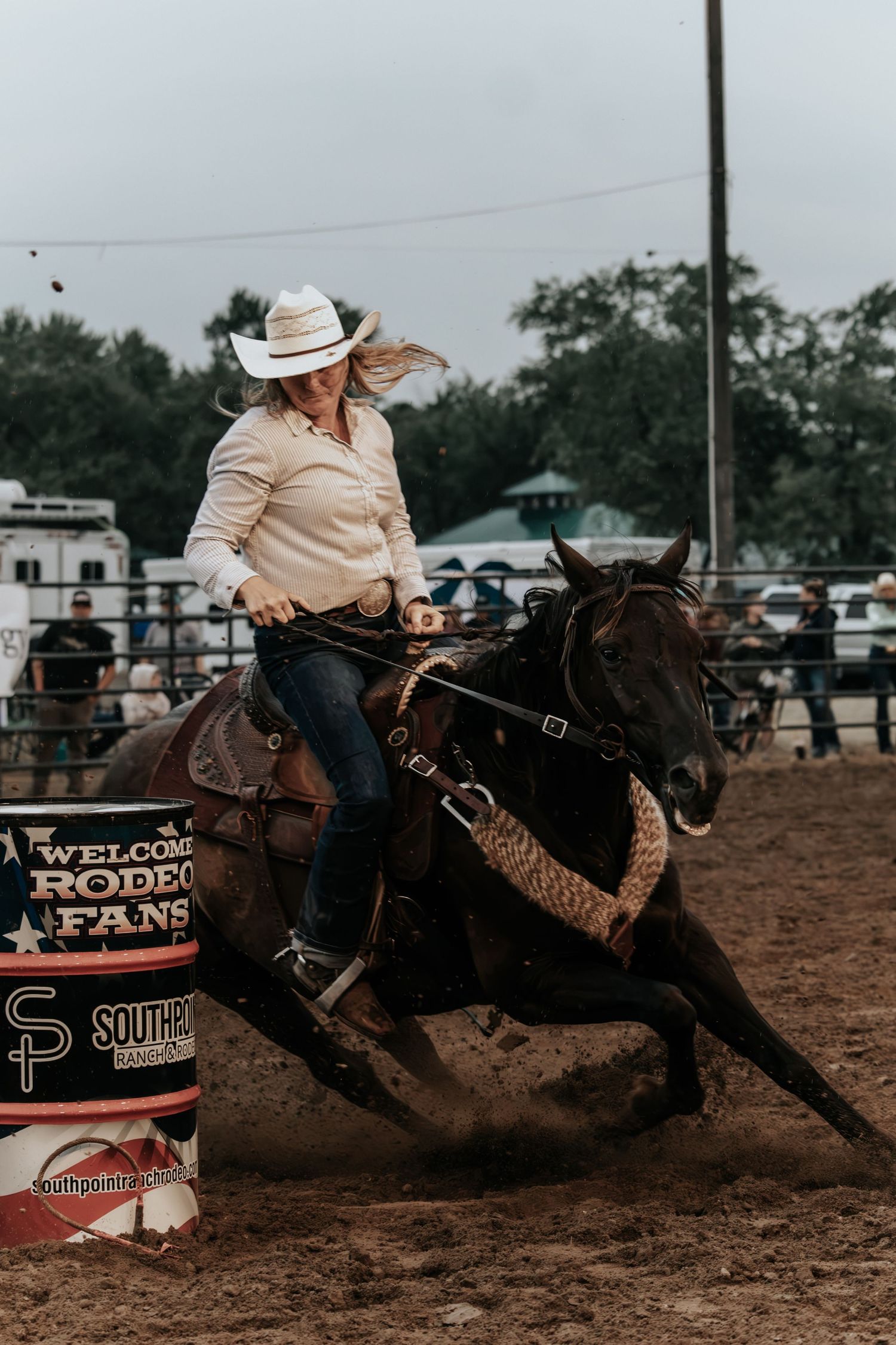 A cowgirl in a white hat rides a dark horse around a barrel at a rodeo competition in a dirt arena.