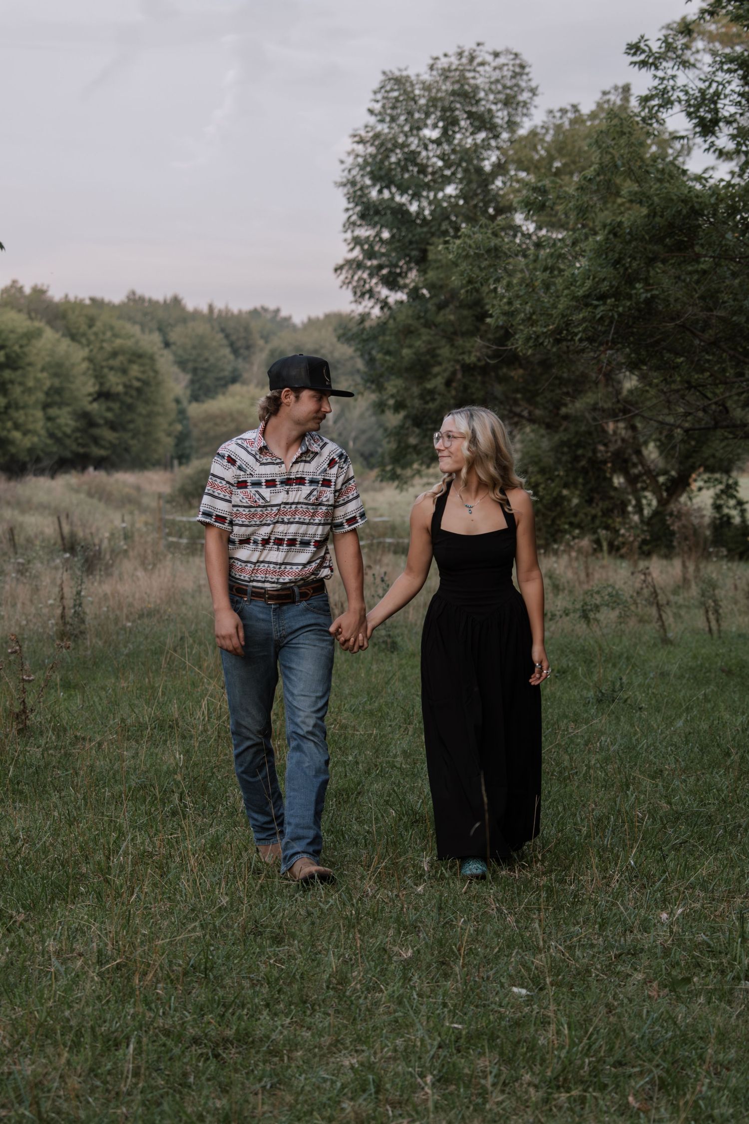 A couple holds hands and walks through a lush green field, smiling at each other on a cloudy day.