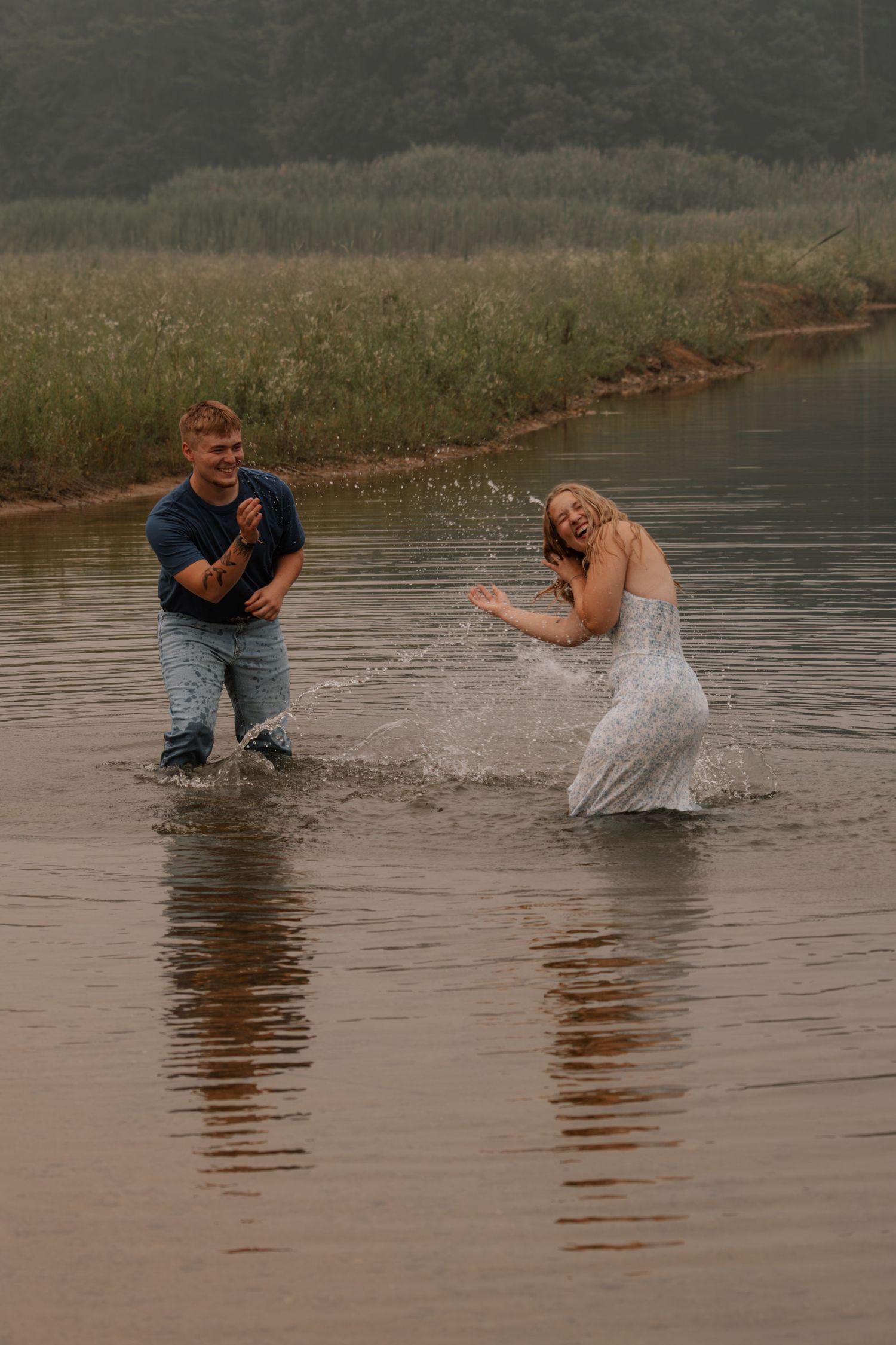 Couple laughing and splashing in a shallow river, woman in white dress and man in jeans enjoying a playful moment.
