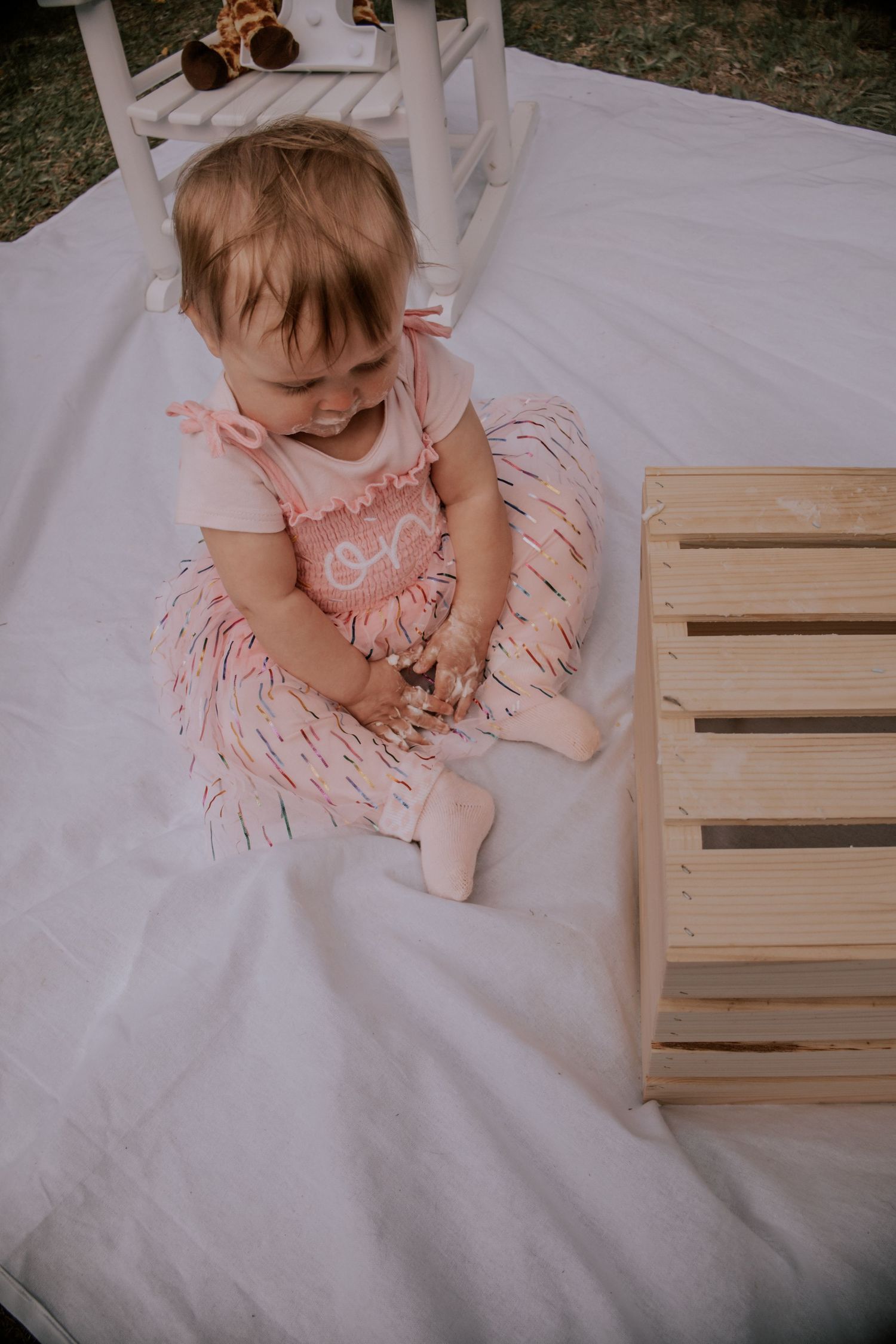 Baby girl in pink outfit sitting on white blanket beside wooden crate at outdoor photo shoot.