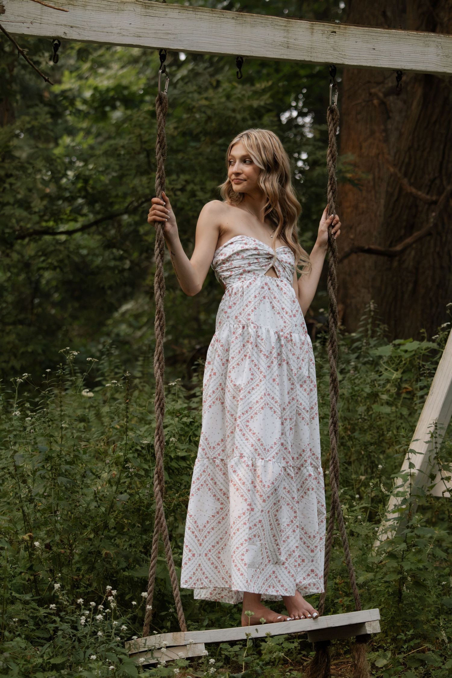 Woman in white patterned strapless maxi dress standing on a wooden swing surrounded by lush green forest.