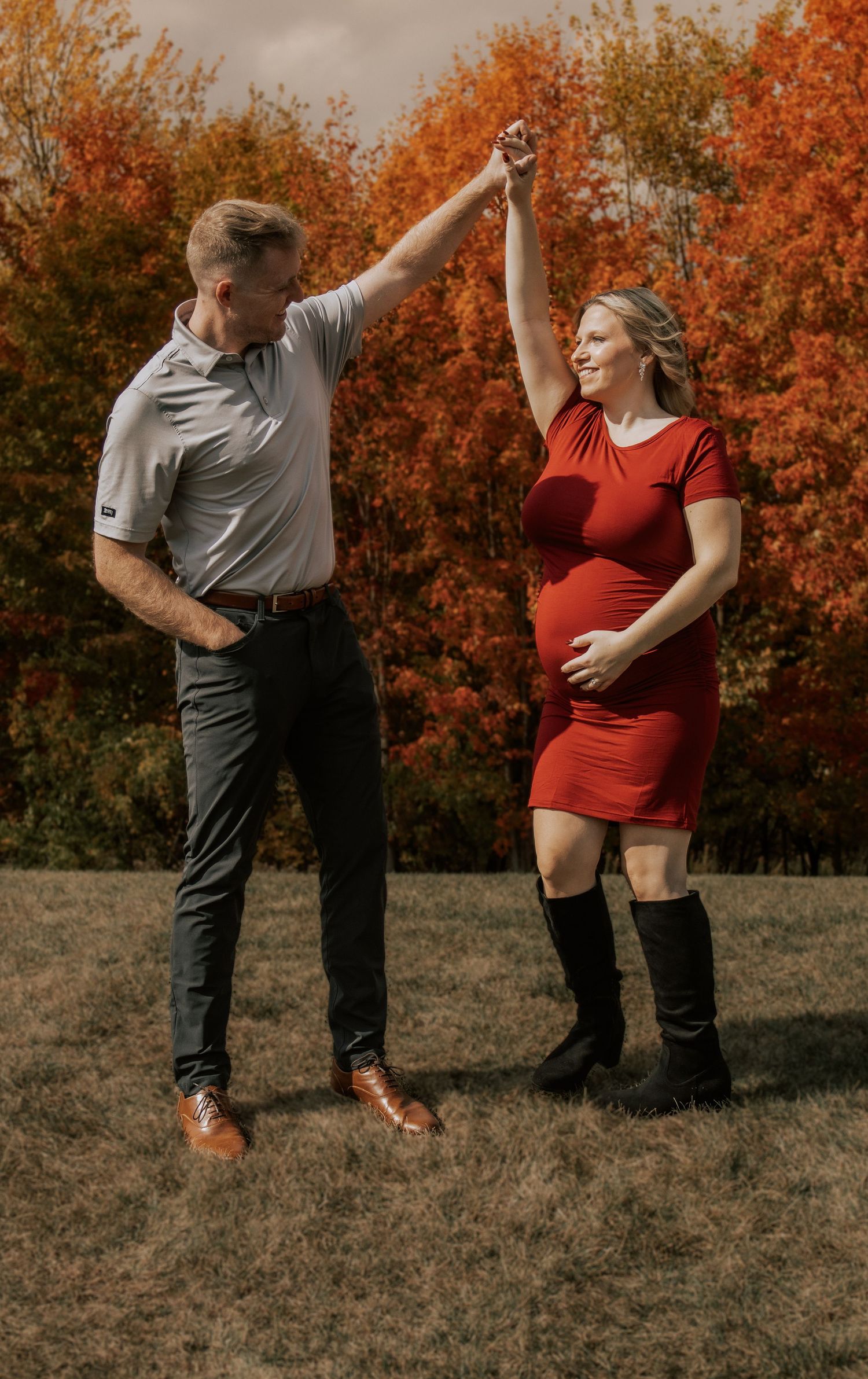 Playful couple dancing together in a field with beautiful orange fall trees creating a warm autumn backdrop.
