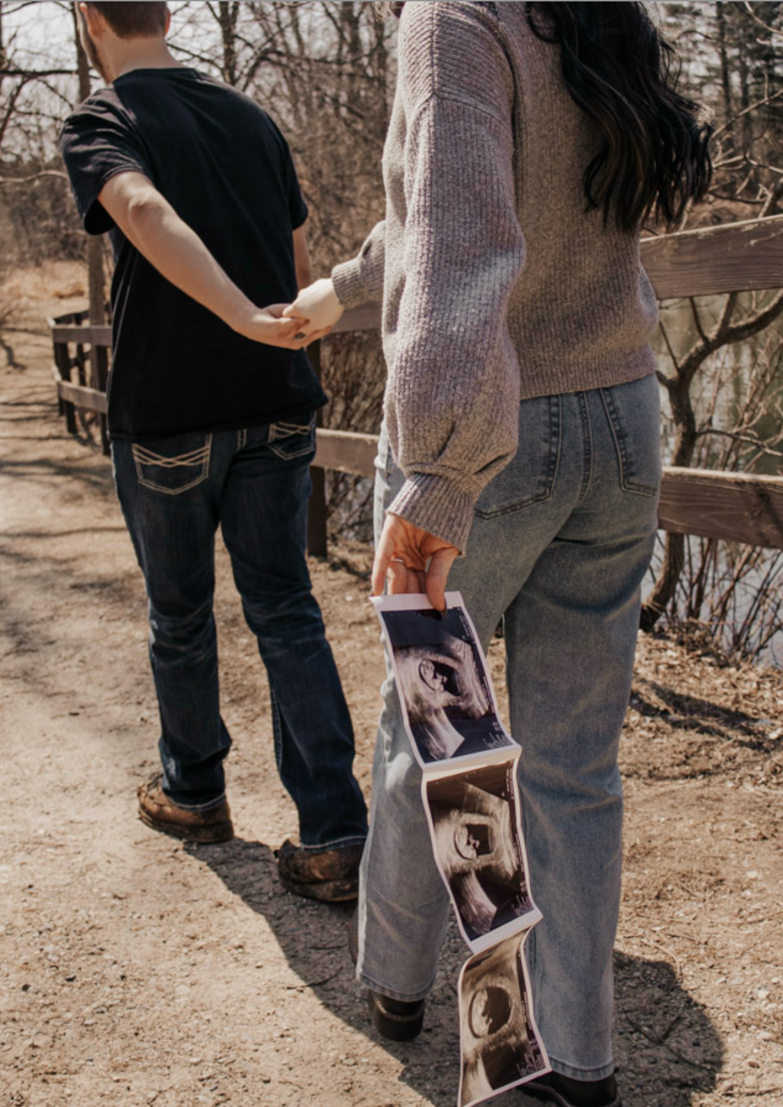 Couple holding hands on a path, woman displaying ultrasound photos, pregnancy announcement outdoors.