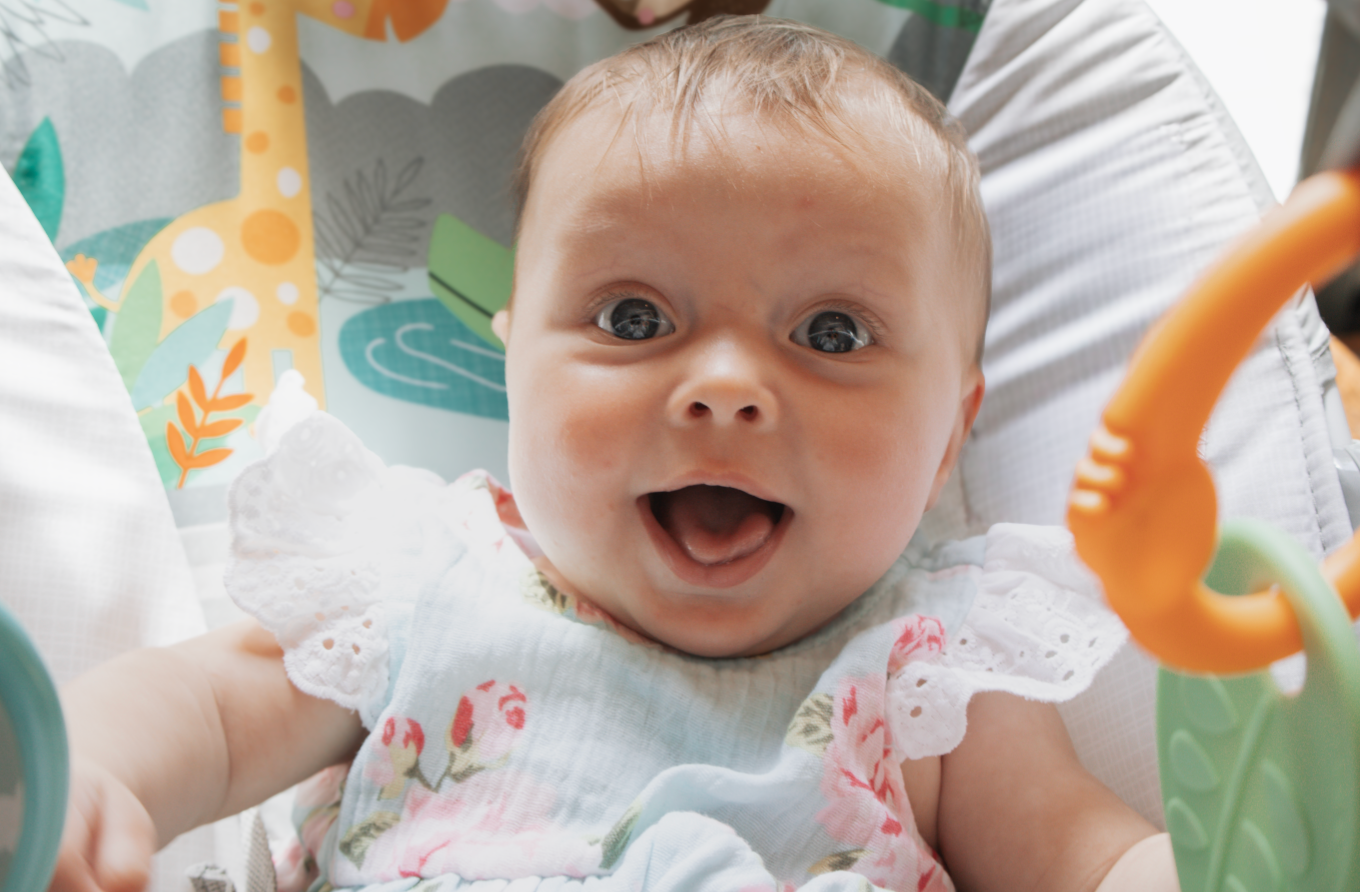 Baby wearing floral dress lies on white bedding with colorful decorative elements in background.