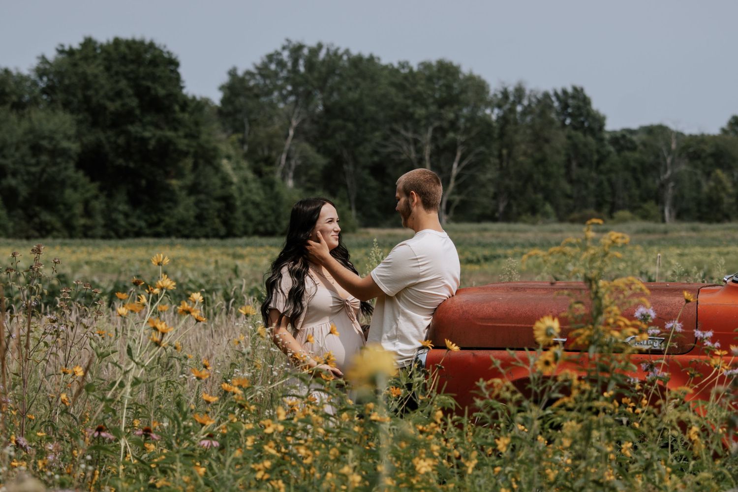 Couple sitting together in a wildflower field near a red tractor, surrounded by lush green trees.