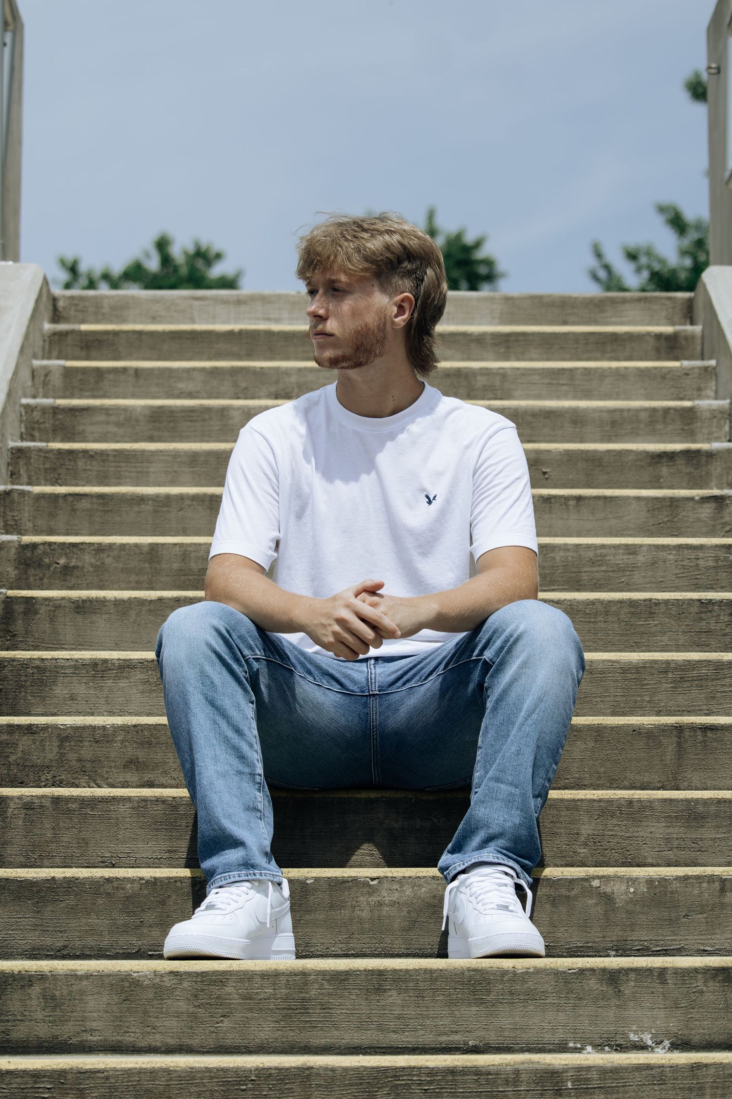 Young man in white t-shirt and jeans sitting on concrete steps outdoors on a sunny day.