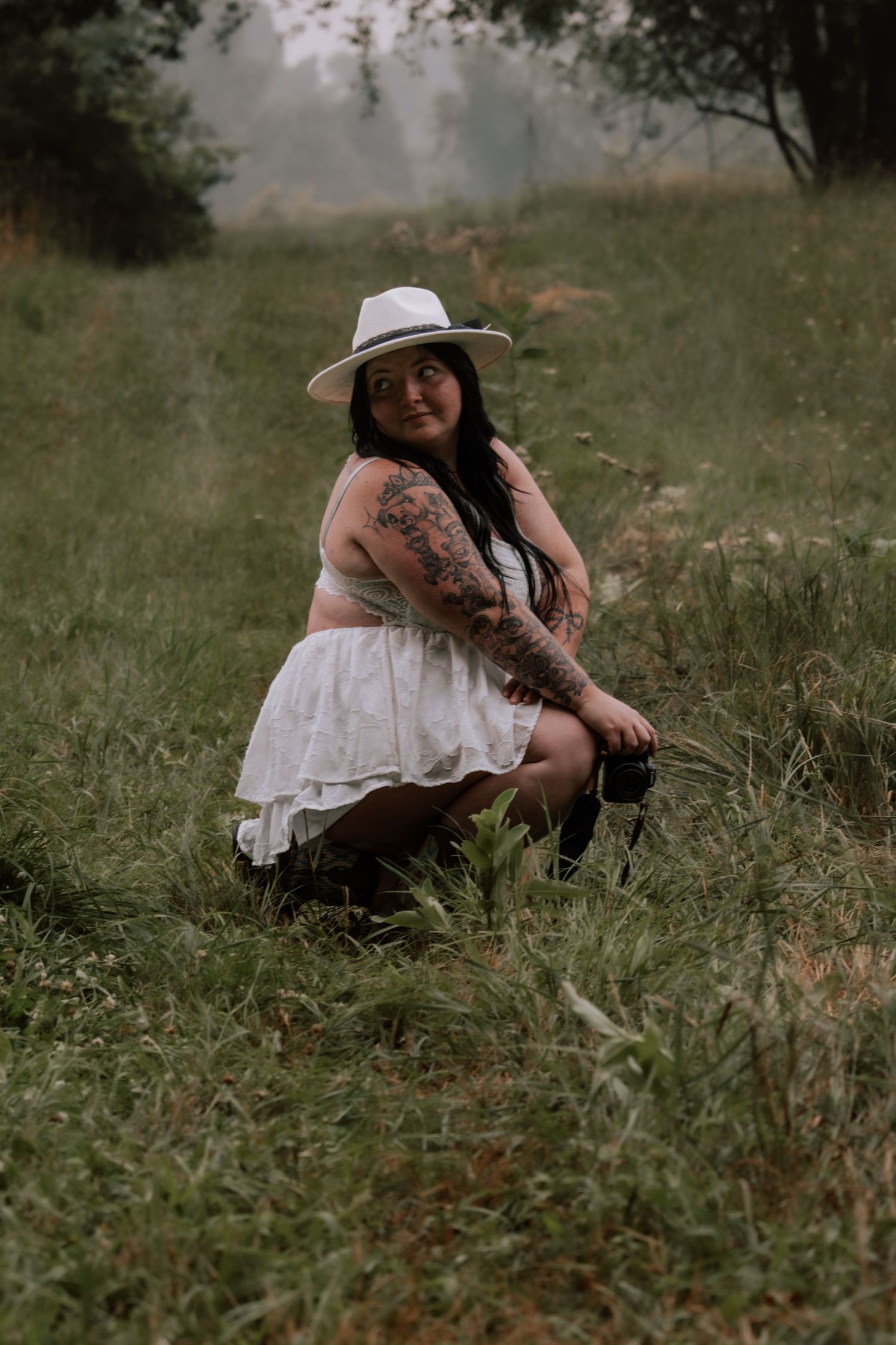 Woman with tattoos wearing white hat and skirt sits in lush green meadow, posing confidently outdoors.