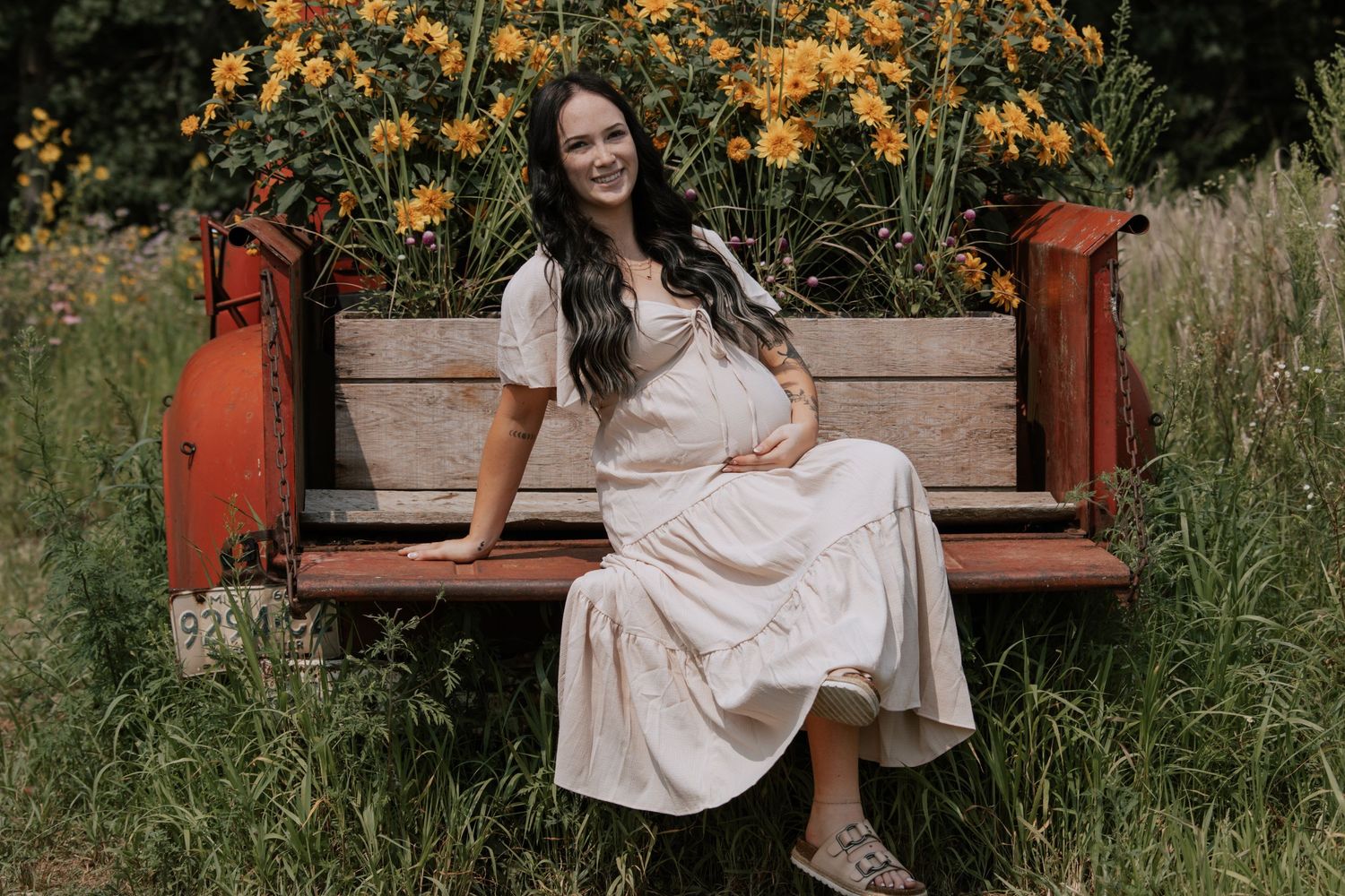 Pregnant woman in white dress sitting on red vintage bench surrounded by yellow wildflowers in a lush garden.