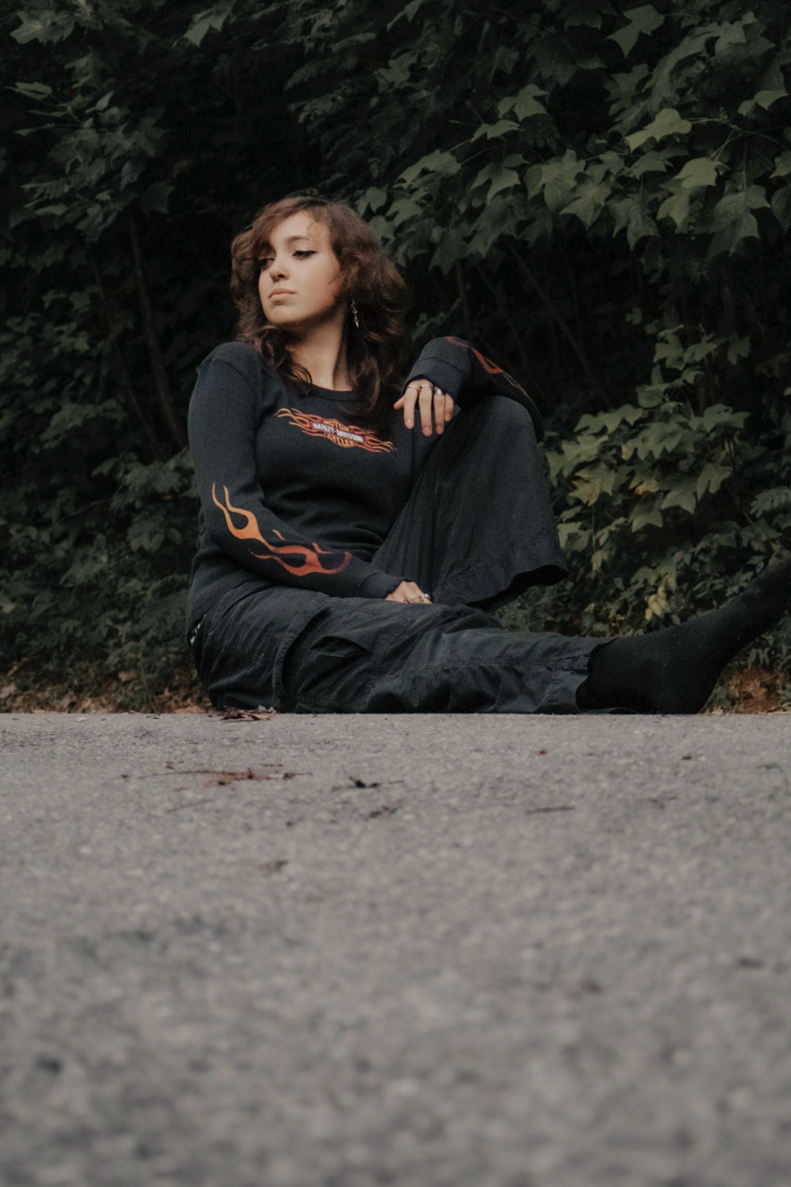 Figure resting on concrete ground surrounded by dark foliage in moody outdoor setting.
