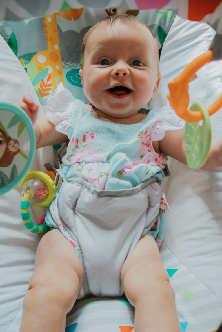 Infant in white and mint outfit smiles while lying on white surface with tropical decorations.