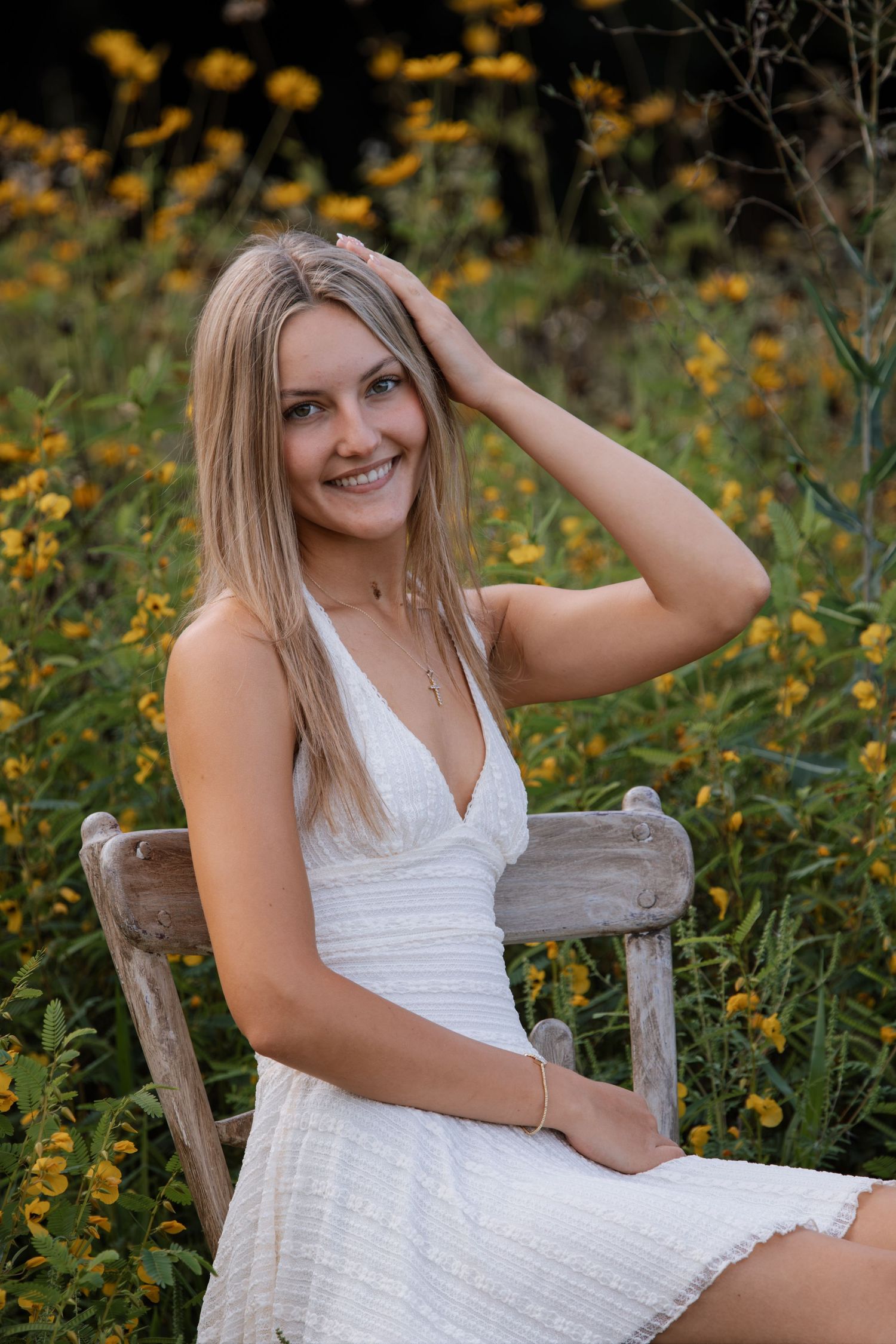 Young blonde woman in white dress sitting on wooden bench surrounded by yellow wildflowers, smiling outdoors.
