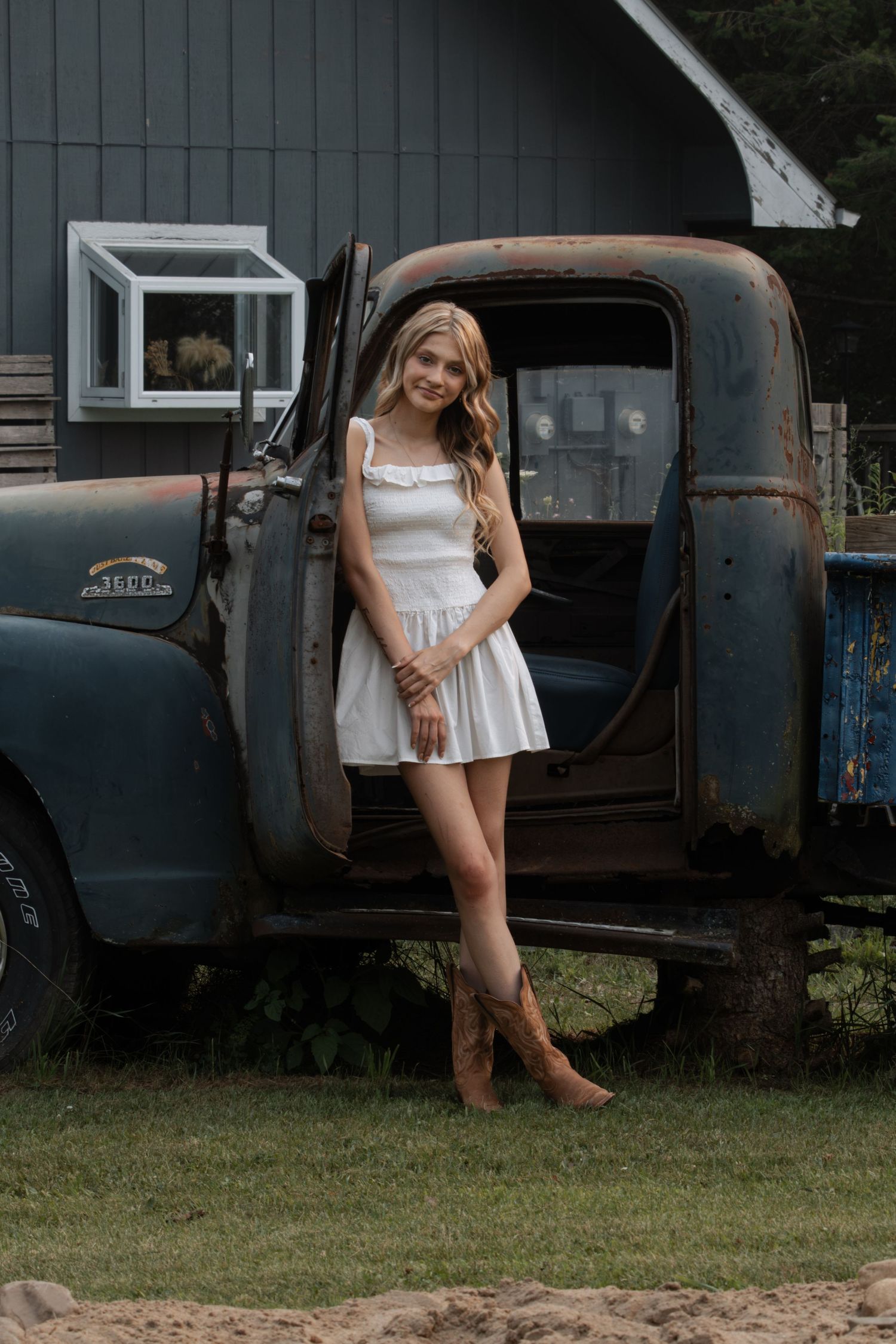 Young woman in white dress and cowboy boots posing beside a vintage rusty truck near a barn.