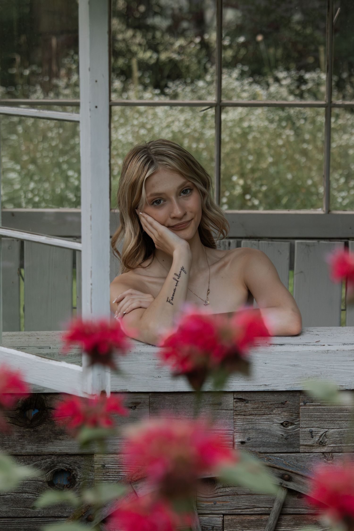 Young blonde woman with tattoos rests her chin on her hand, smiling softly by a rustic window with red flowers.