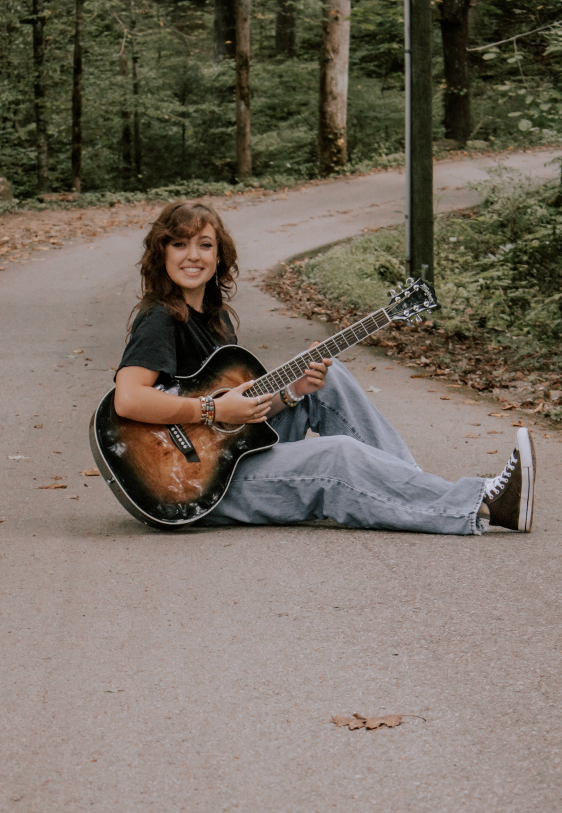 A musician sitting on a curved path in the woods playing an acoustic guitar while wearing casual clothes and sneakers.