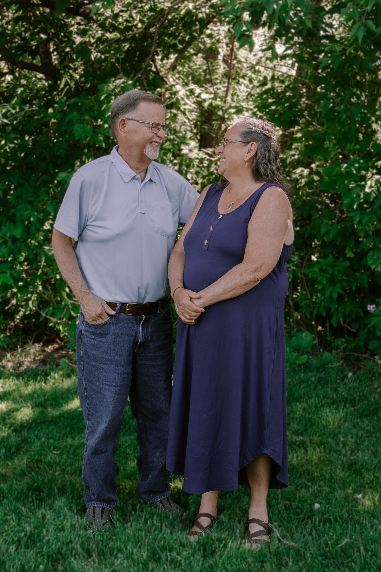 Older couple smiling at each other outdoors, man in blue polo and jeans, woman in navy dress, green trees background.