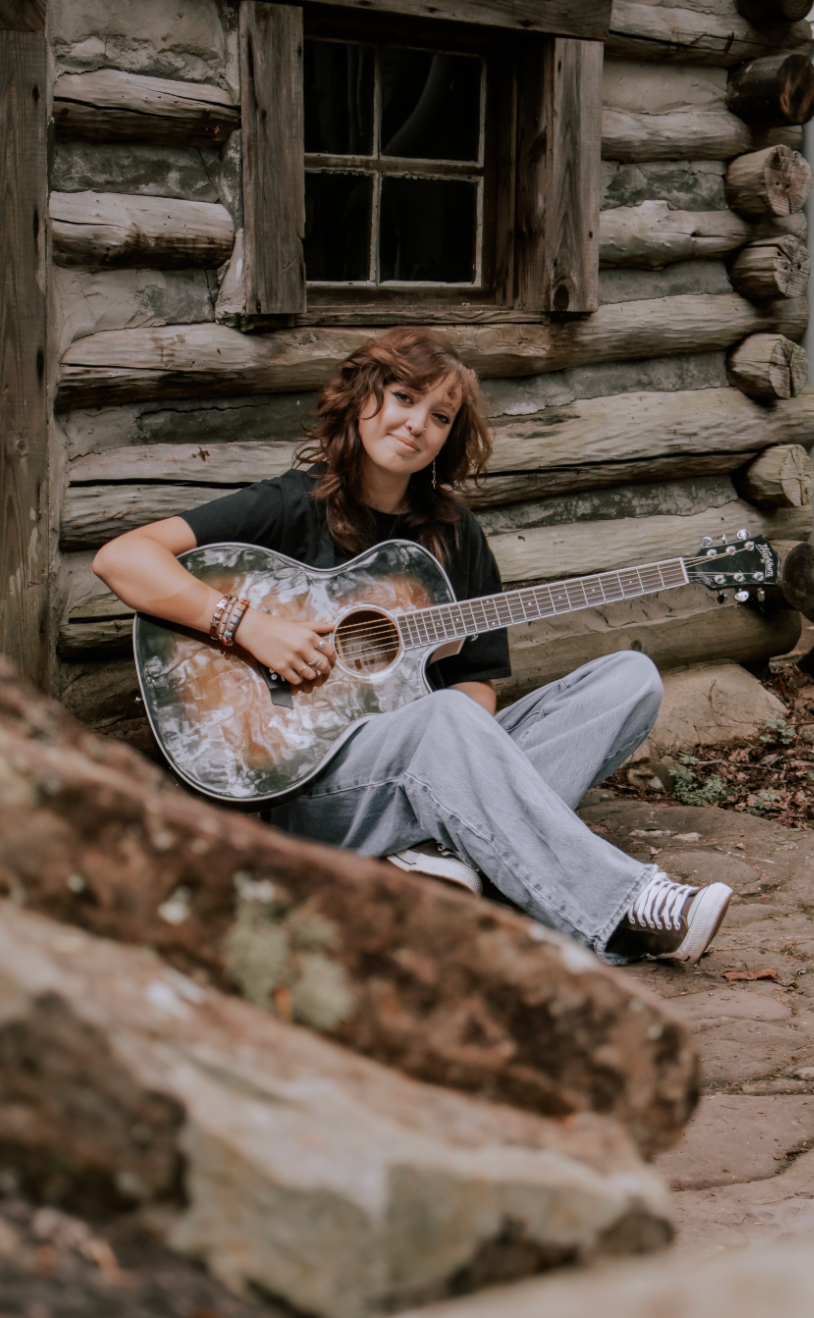 Young woman playing acoustic guitar while sitting against a rustic log cabin wall outdoors.