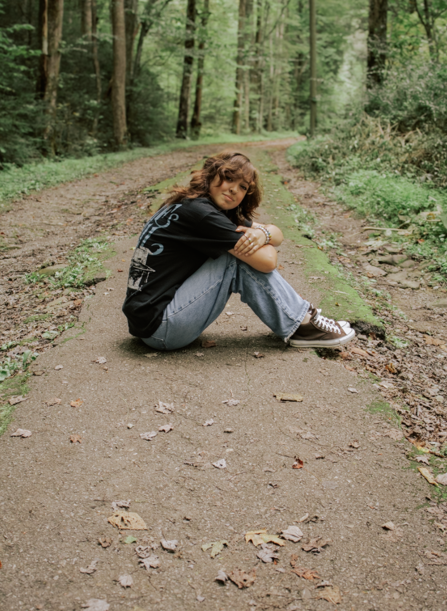 Someone rests on a dirt path in a lush green forest while holding a guitar and backpack.