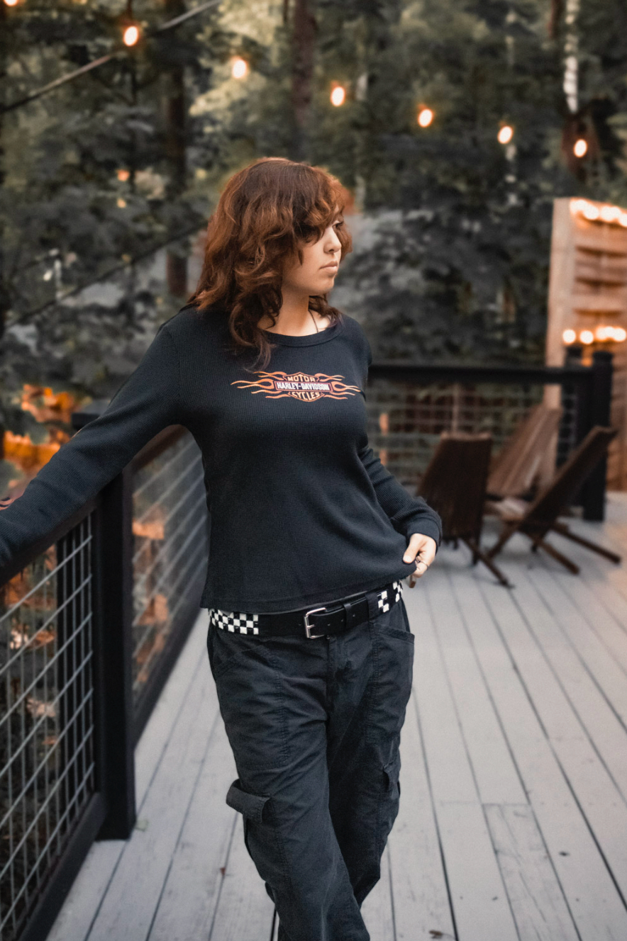 Person in black sweater and checkered belt walking on wooden deck with string lights at sunset.