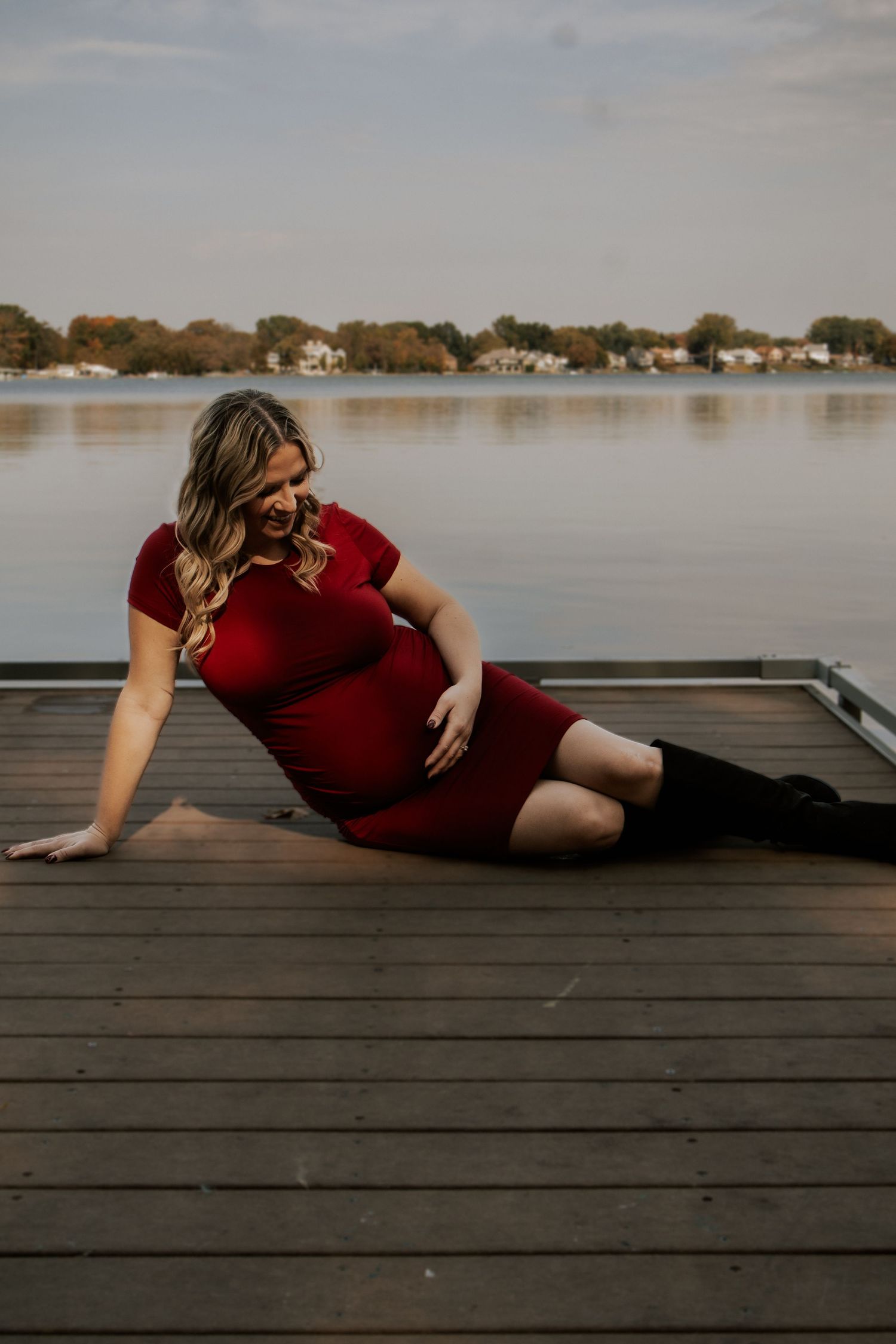 Artistic maternity pose on wooden dock with lake and houses in background.