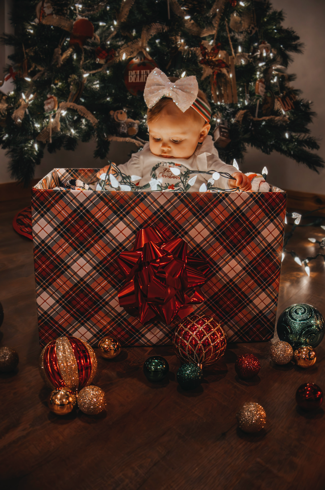 Christmas presents wrapped in plaid paper with red bows sit beneath a decorated tree with ornaments and lights.