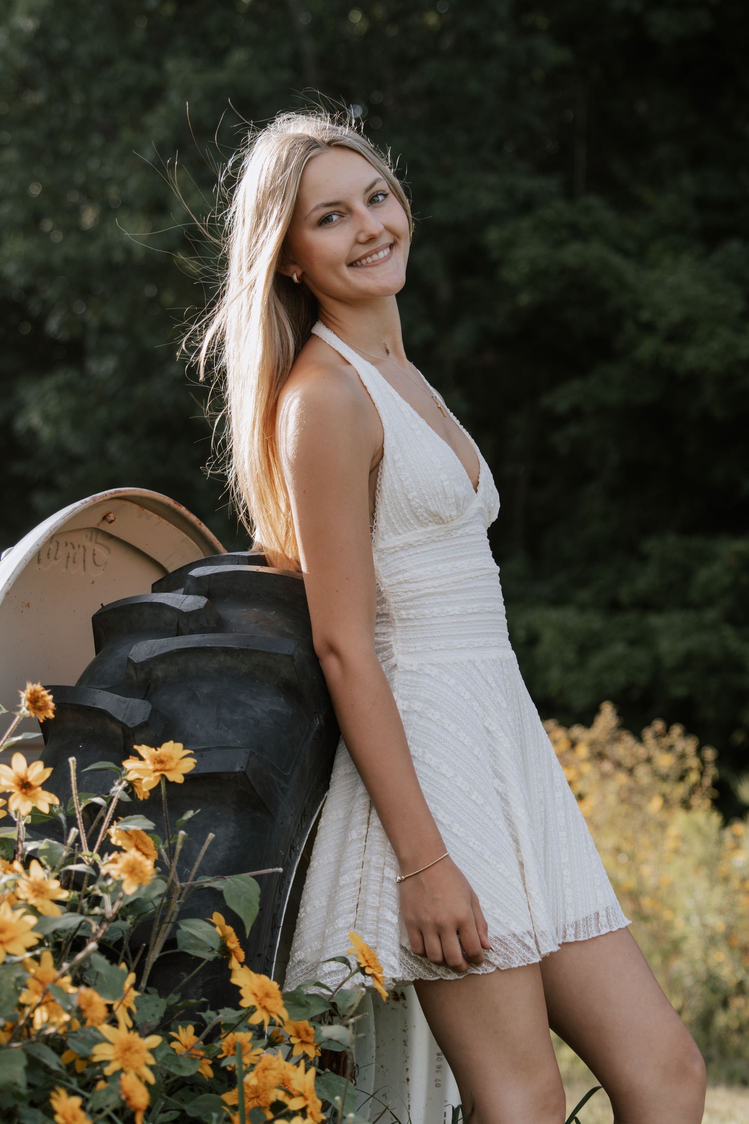 Young blonde woman in white sundress smiling outdoors near yellow flowers and a tractor tire.