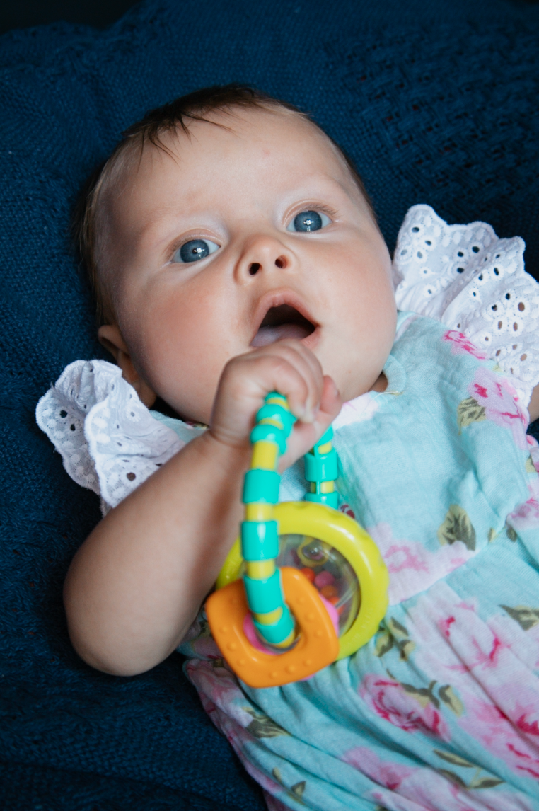 Baby with teething toy lies on dark blue background wearing lace-trimmed outfit.