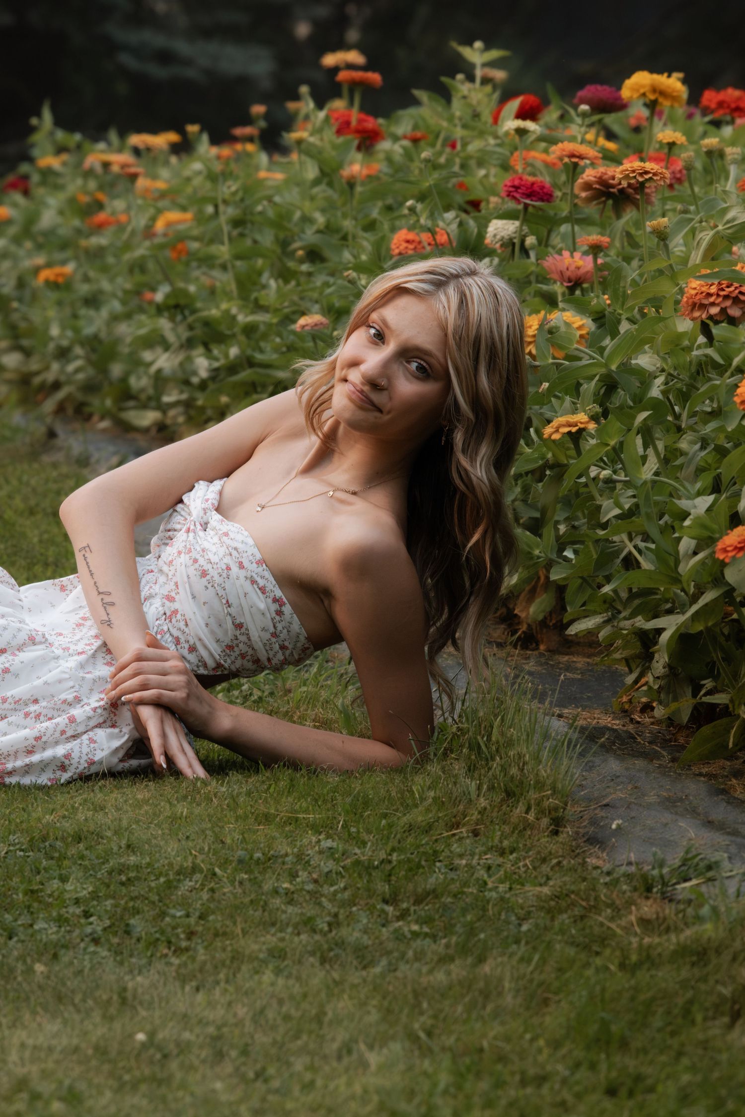 Young woman in floral dress reclining on grass beside colorful zinnia garden flowers outdoors.