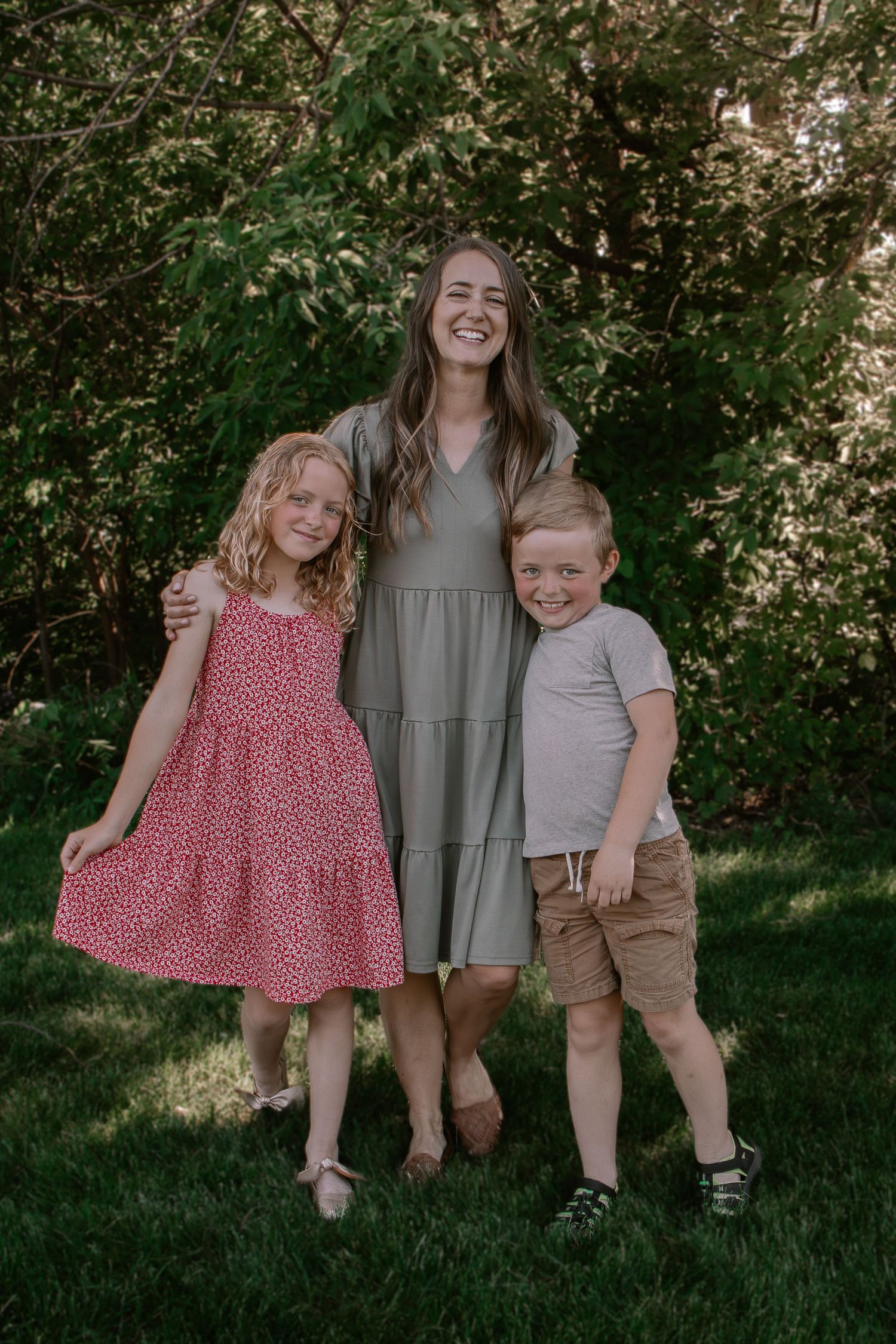 A smiling woman in a sage dress poses outdoors with two young children, a girl in red and a boy in gray.