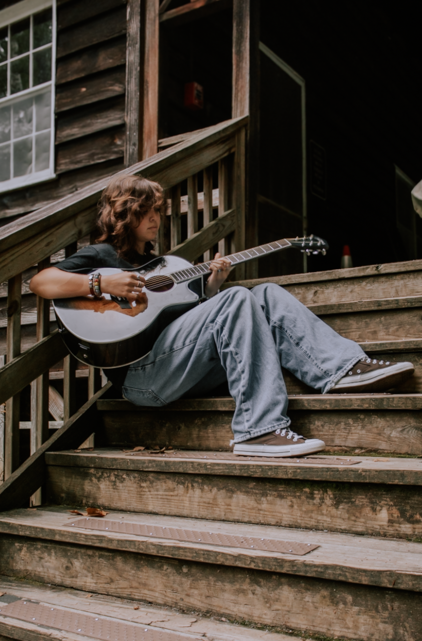 A person relaxes on wooden steps with an acoustic guitar, showcasing casual style in denim and sneakers.