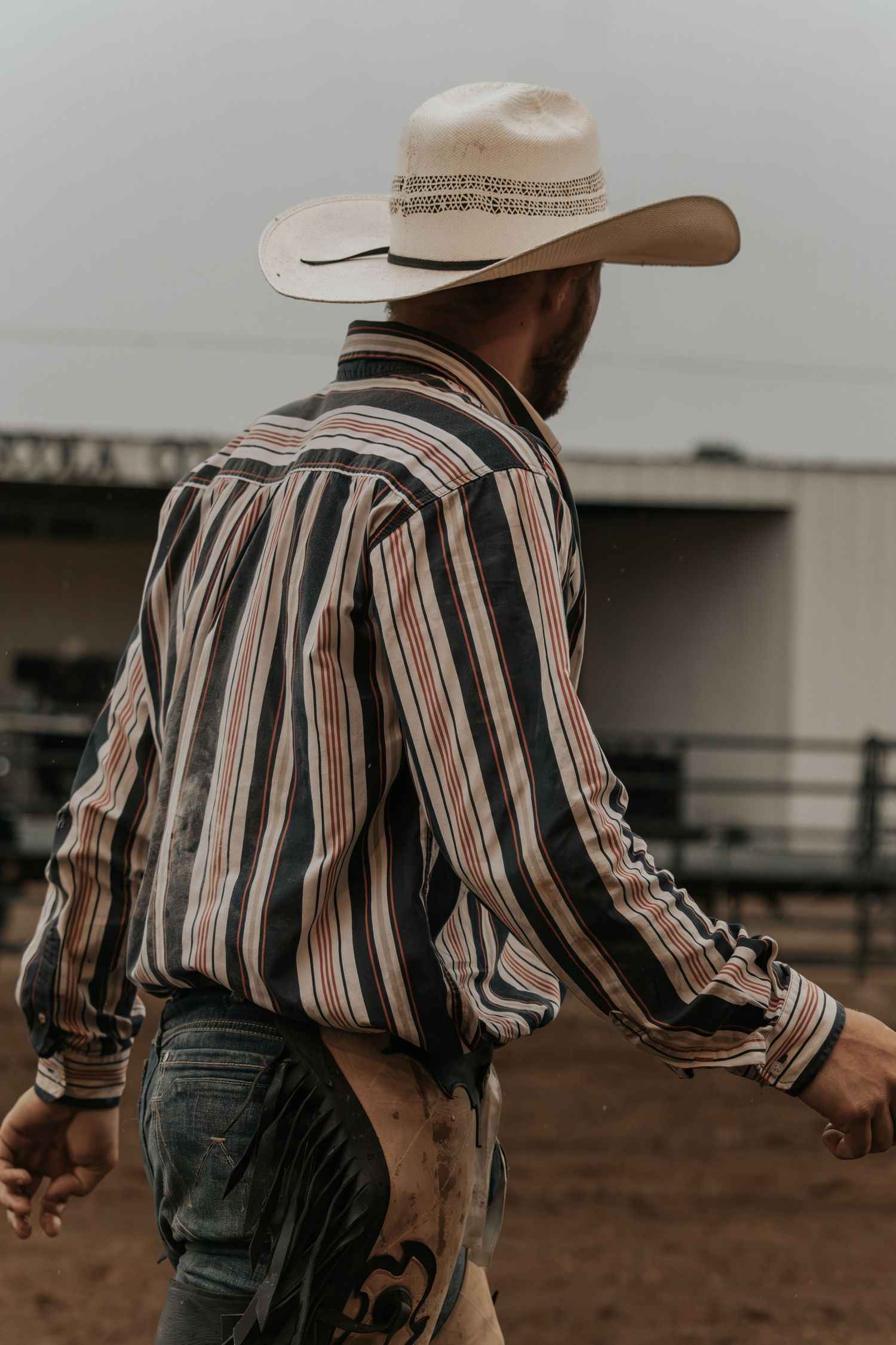 A cowboy in a striped western shirt and white hat walks through a dusty rodeo arena.