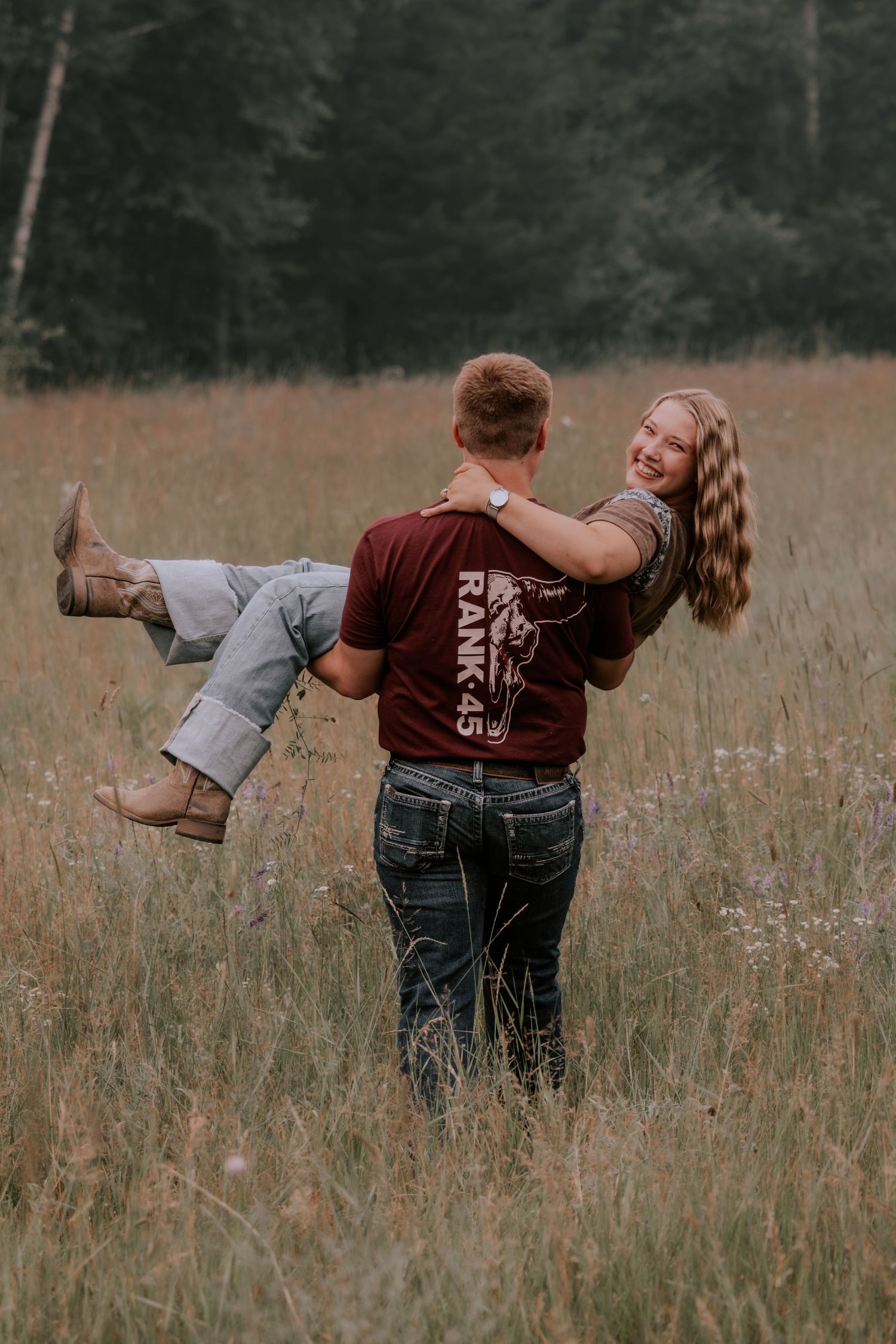 A man lifts a smiling woman in a sunlit meadow, both wearing casual western attire with tall grass and trees behind them.