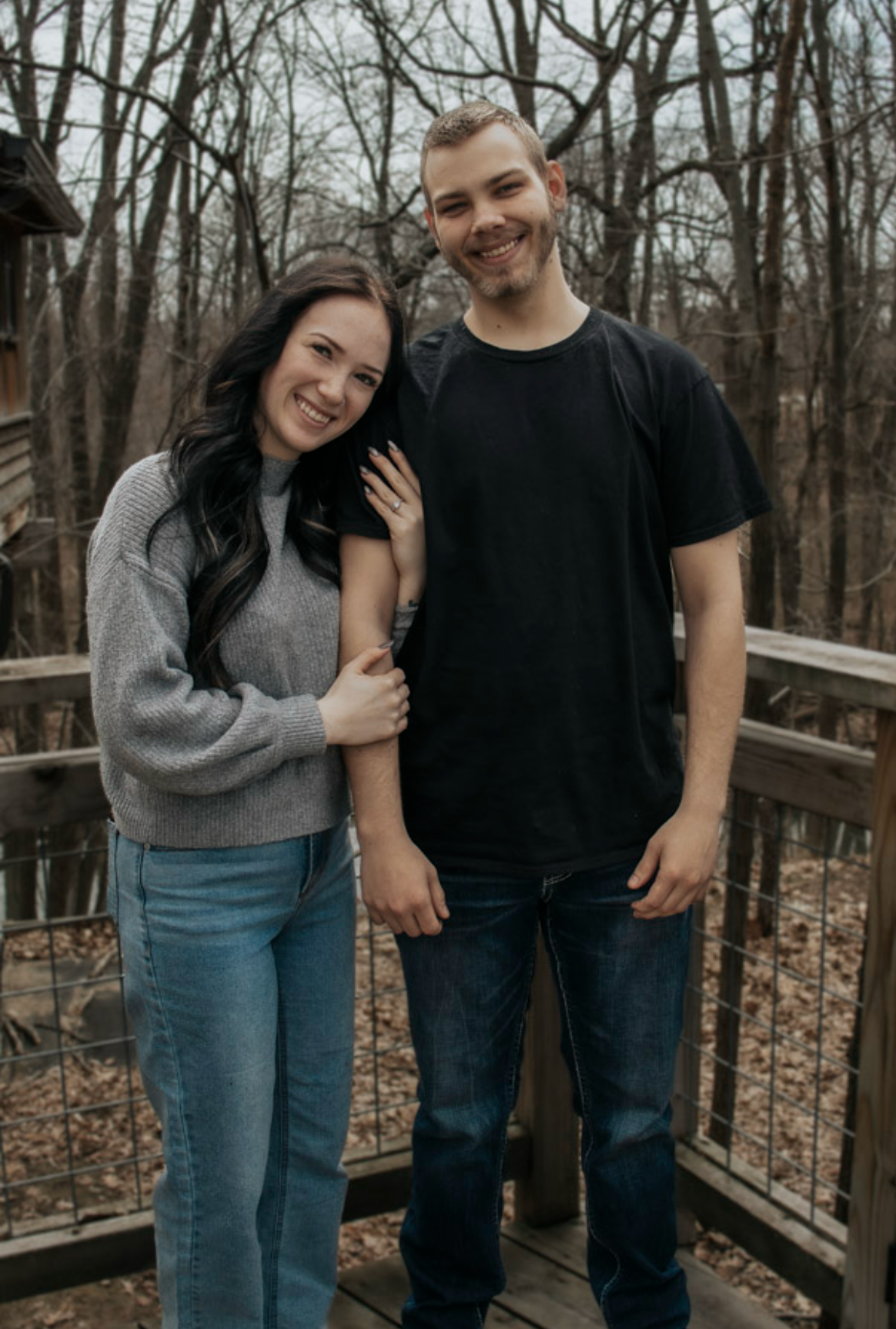 A smiling couple poses on a wooden deck surrounded by bare trees, the woman in a gray sweater and the man in a black t-shirt.