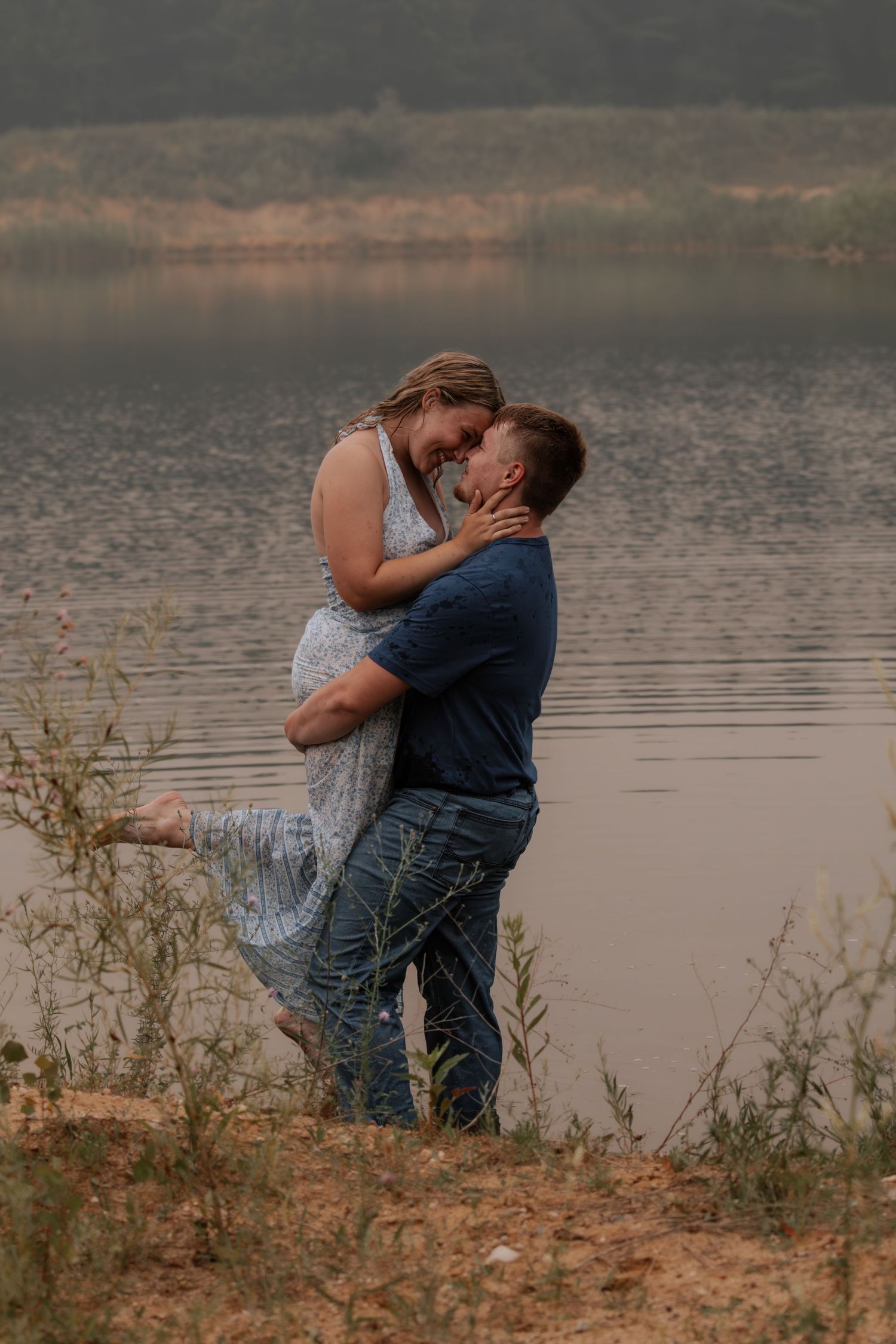 Couple embracing romantically by a misty lake, man lifting woman in floral dress near sandy shore with lush greenery.