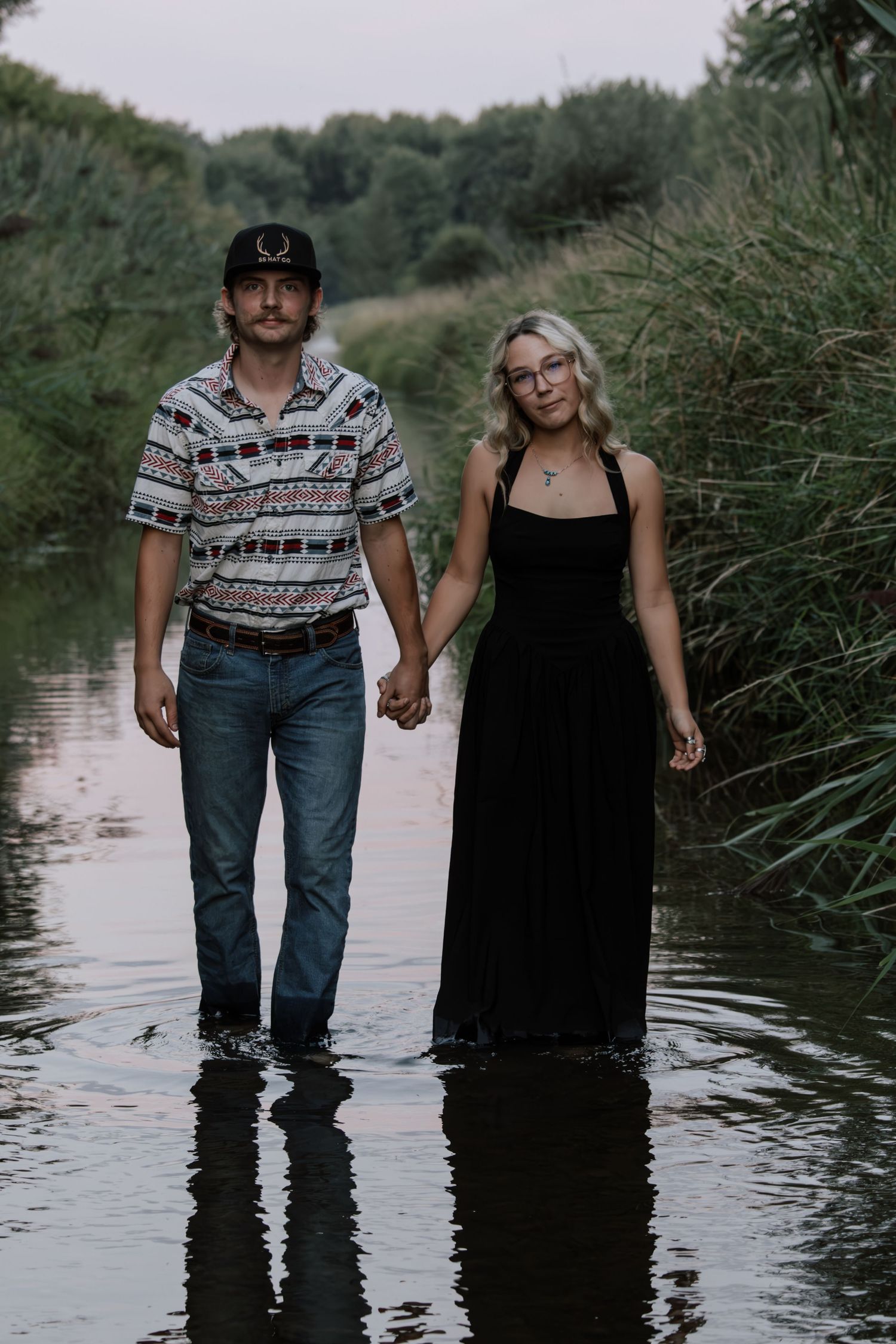 Couple holding hands standing in shallow water surrounded by lush greenery, woman in black dress, man in patterned shirt.